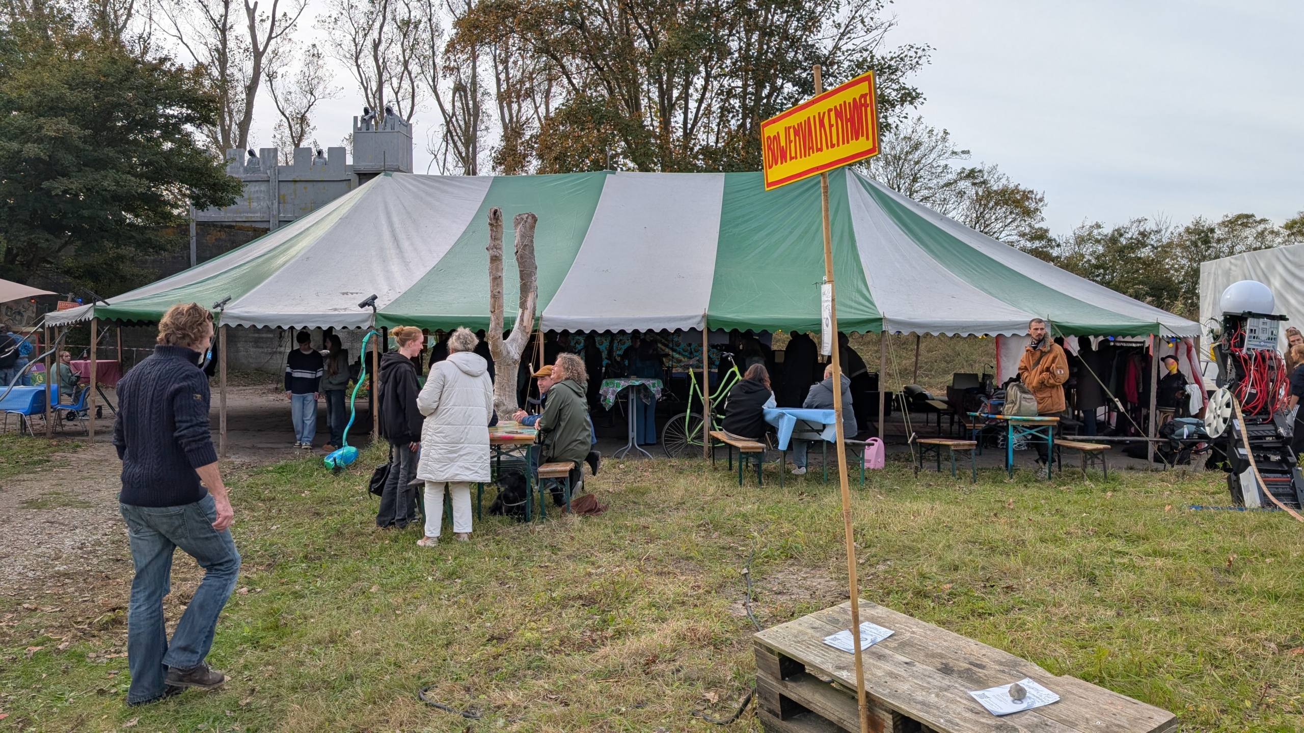 Een groen-wit gestreepte tent met mensen ervoor en erin tijdens een evenement. Een geel-rood bord met de tekst 'BOUWENWALKENNAAR' staat in de voorgrond. In de tent zitten en staan mensen, en er is een persoon met machines aan de rechterkant. Op de achtergrond staan bomen en een kasteelachtig gebouw.