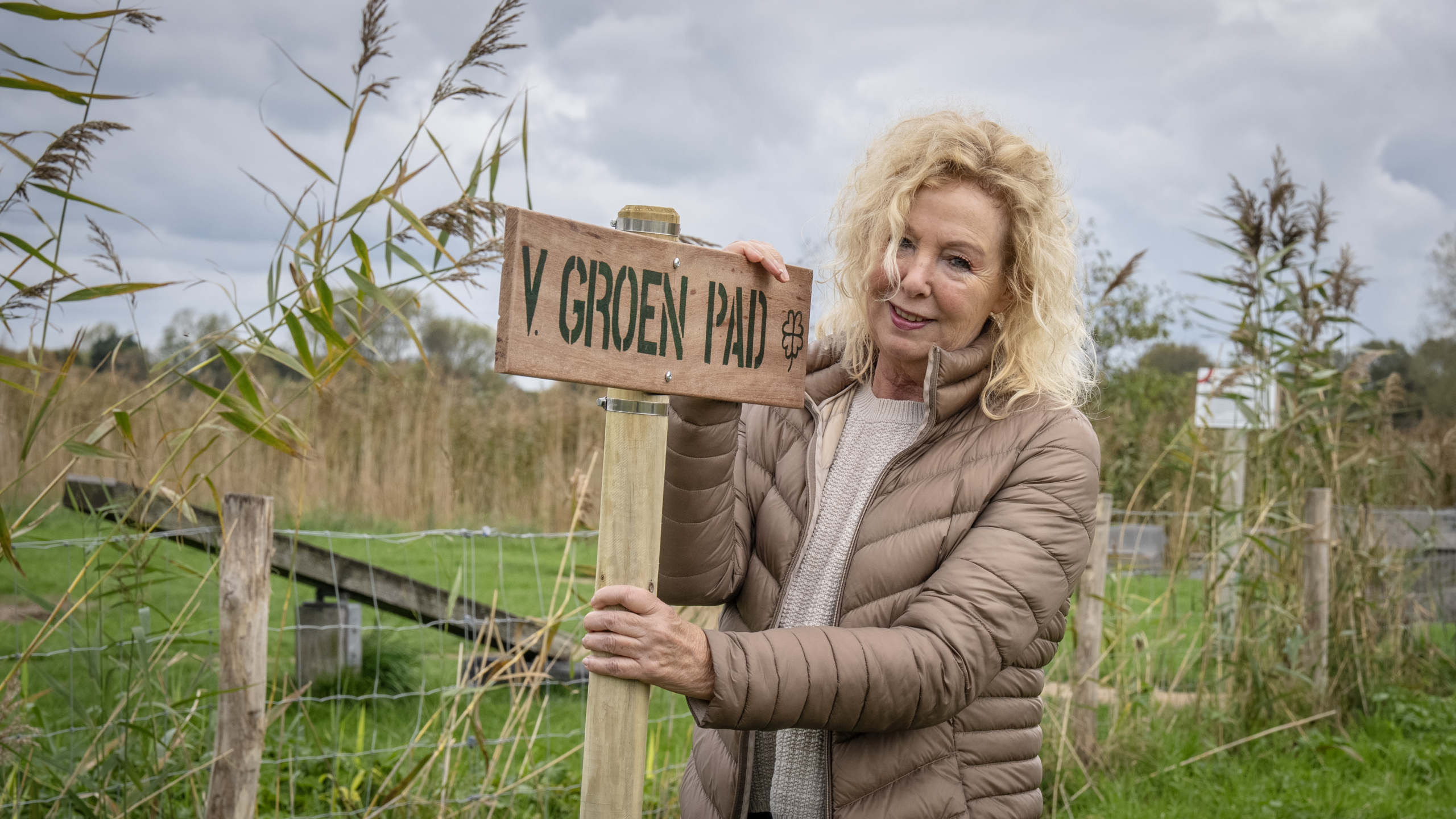 Vrouw met blond haar en een beige jas staat naast een houten bord met de tekst 'V. GROEN PAD' in een groen landschap met hoge grassen.