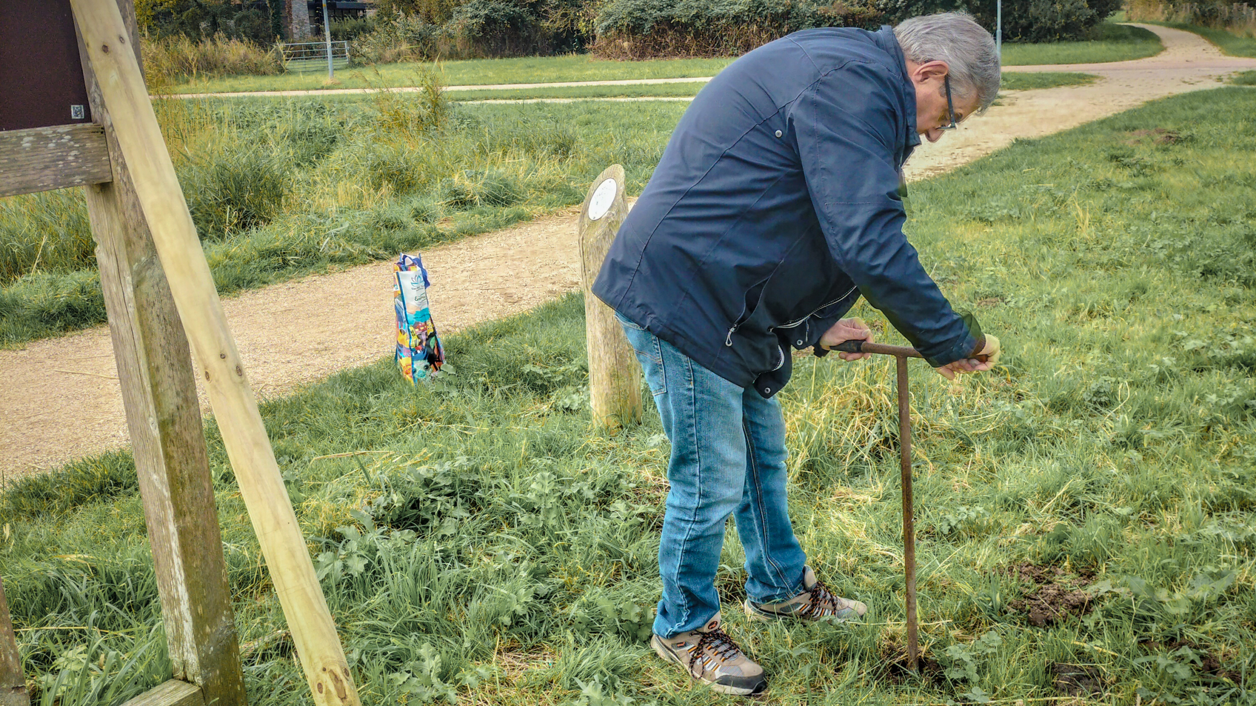Een man buigt voorover op gras met een metalen staaf in de grond, omringd door een parkachtige omgeving.