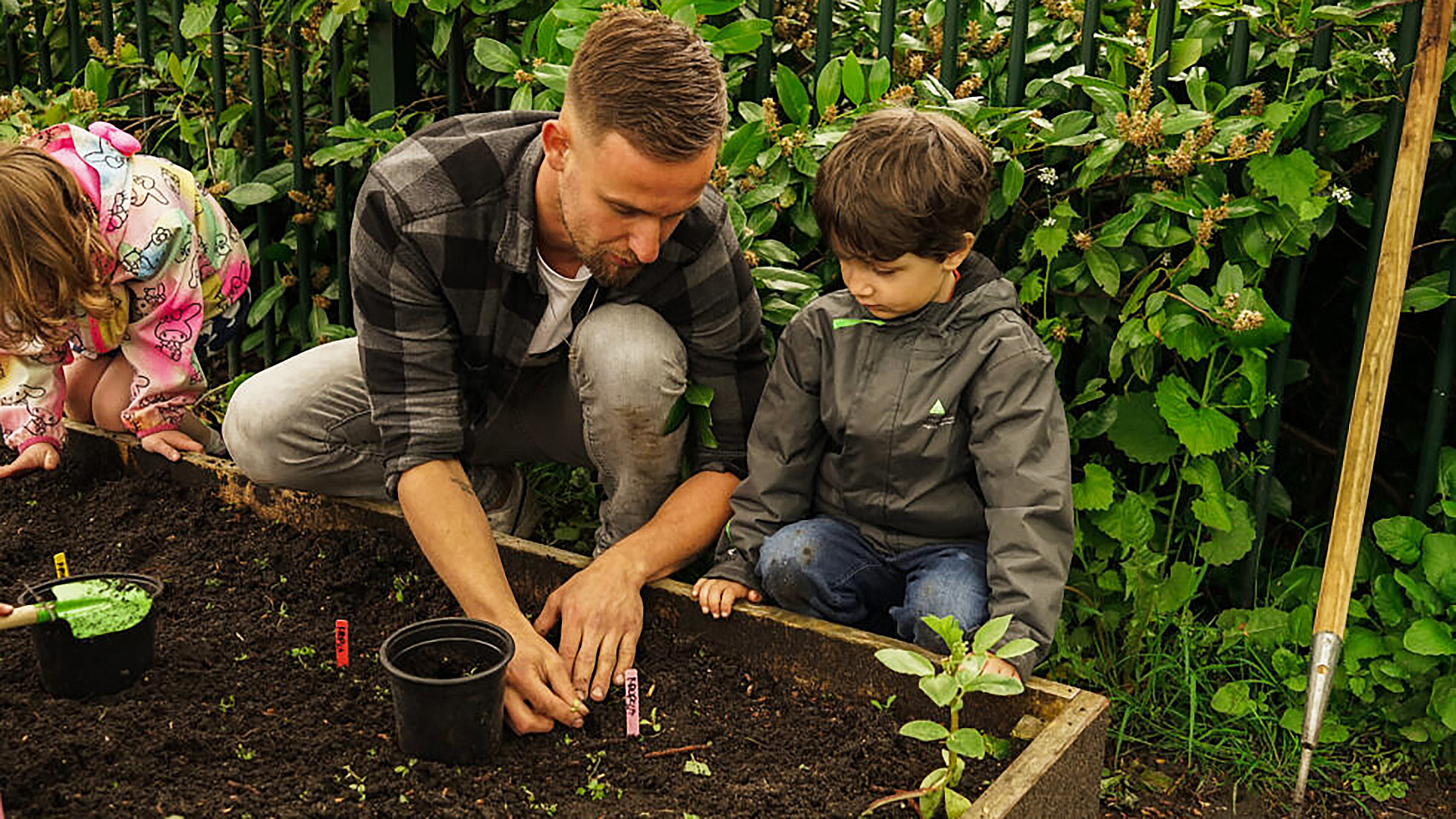 Een volwassene en twee kinderen planten zaadjes in een tuinperceel, omringd door groene struiken.