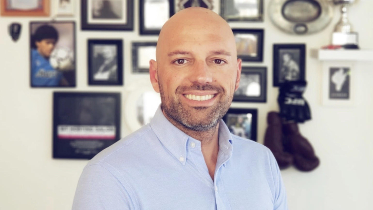 Een kale man met een baard in een lichtblauw overhemd staat voor een muur met ingelijste foto's en trofeeën.