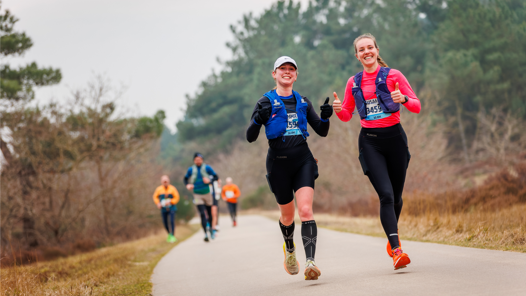 Twee hardlopers, een in een roze shirt en de ander in een zwarte outfit met een pet, rennen zij aan zij op een landweg en geven een duim omhoog; op de achtergrond rennen andere mensen in een natuurlijke omgeving met bomen.