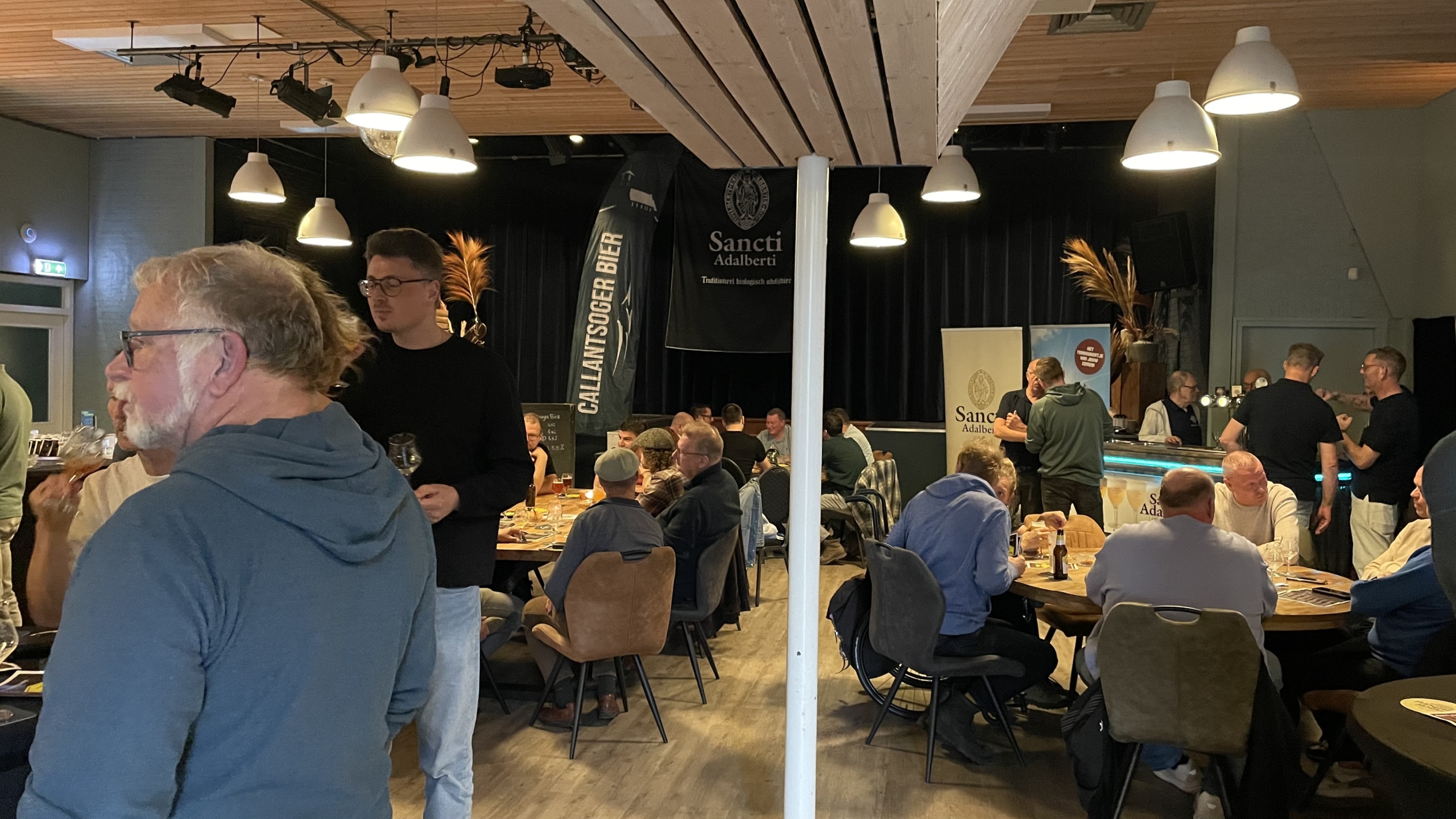 Een groep mensen verzameld in een café-achtige ruimte met tafels en stoelen; enkele mensen staan bij de bar, terwijl anderen aan tafels zitten en met elkaar praten. Er zijn banners met de tekst 'Sancti Adalberti'.