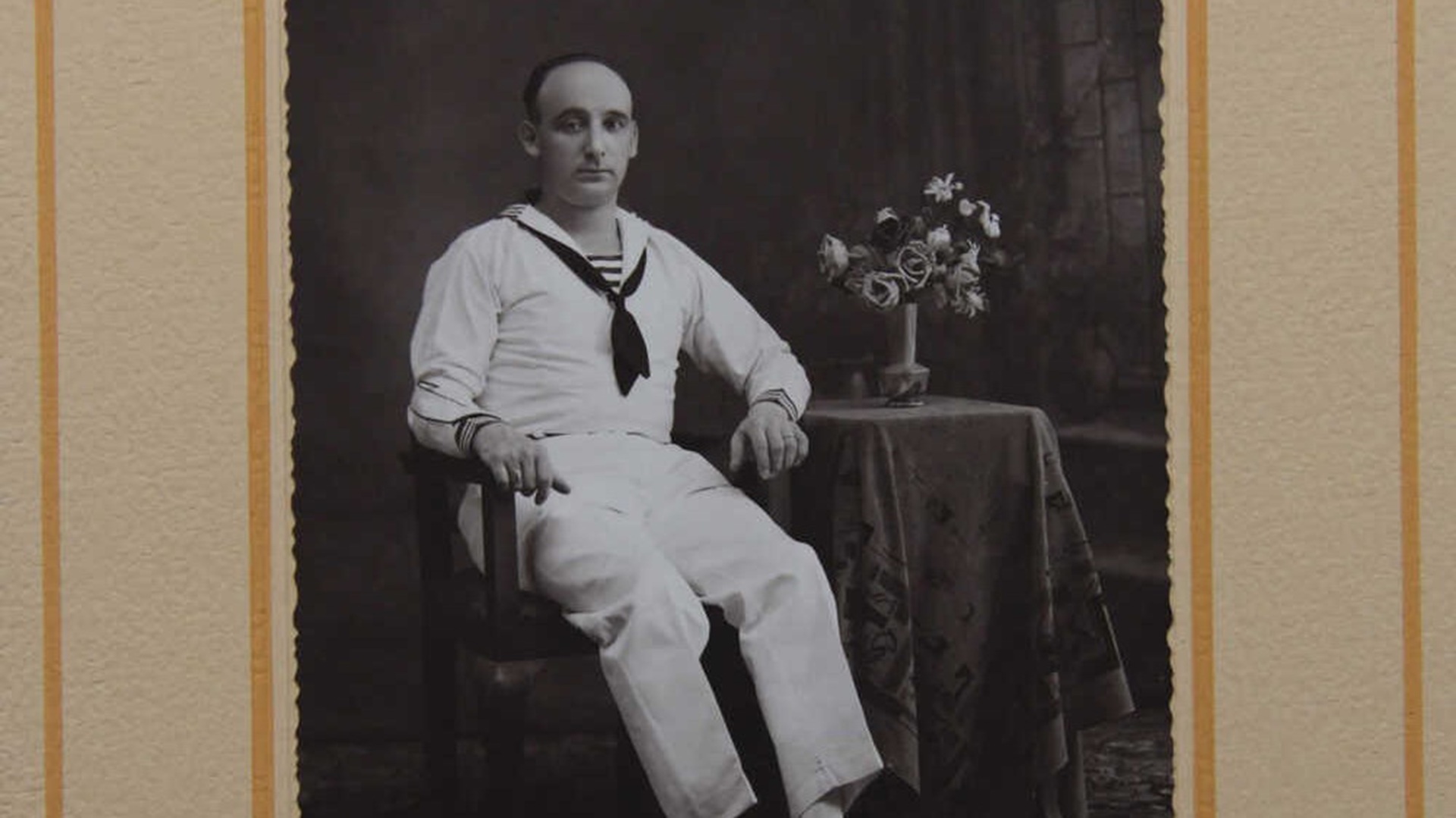 Zwart-wit foto van een man in een matrozenuniform die op een stoel zit naast een tafeltje met bloemen in een vaas.