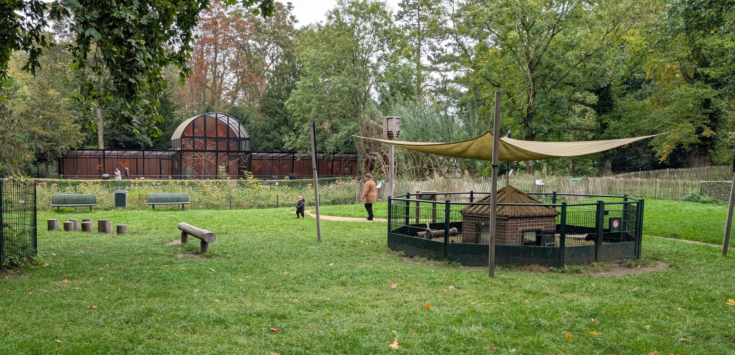 Een speeltuin met grasveld, houten speeltoestellen, en een omheind klein dierverblijf met een doek als zonnescherm, omringd door bomen en een groot gebouw op de achtergrond.
