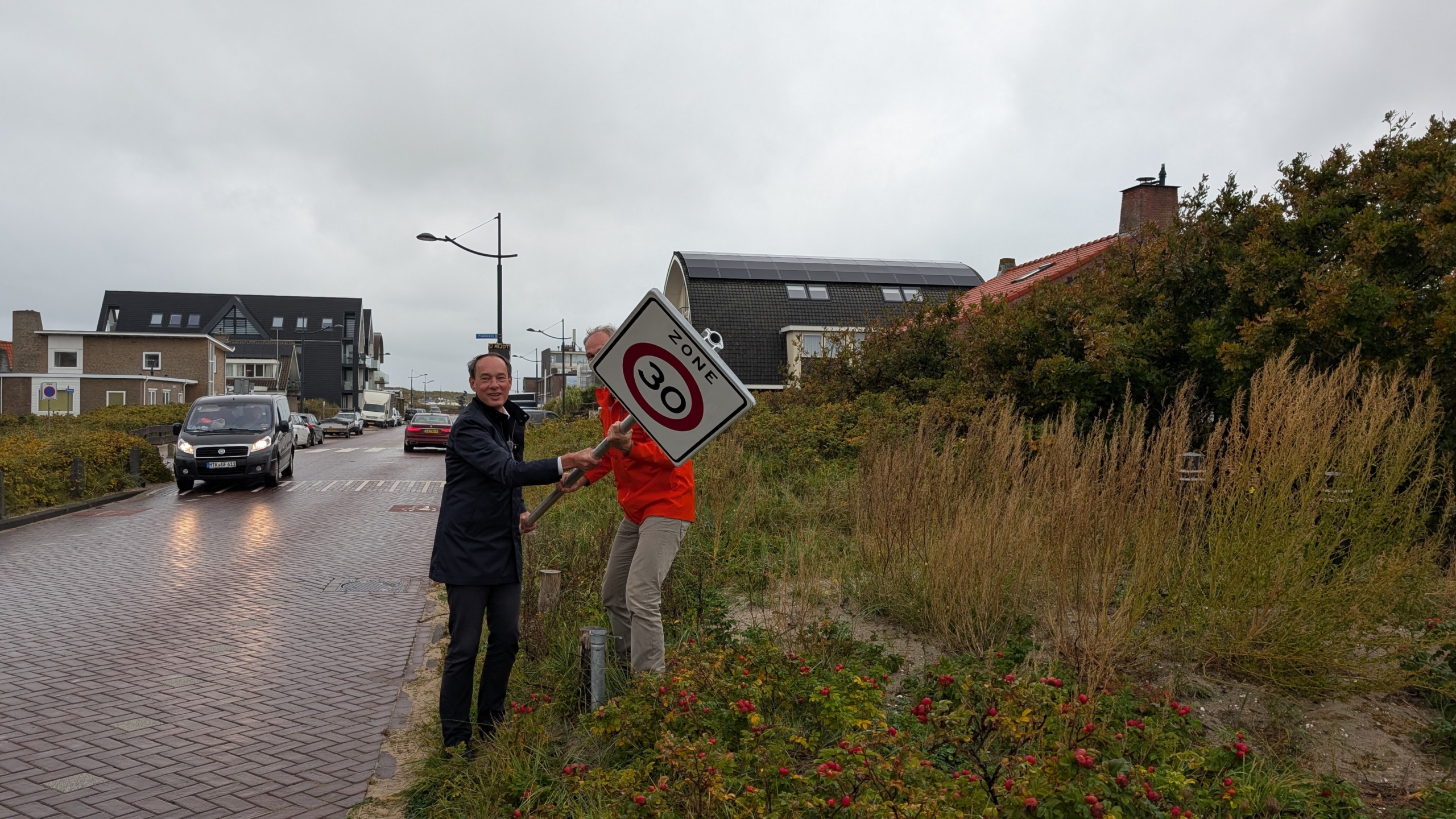 Twee mannen plaatsen een verkeersbord met een snelheidslimiet van 30 km/u langs een weg in een woonwijk met bakstenen huizen en struiken.