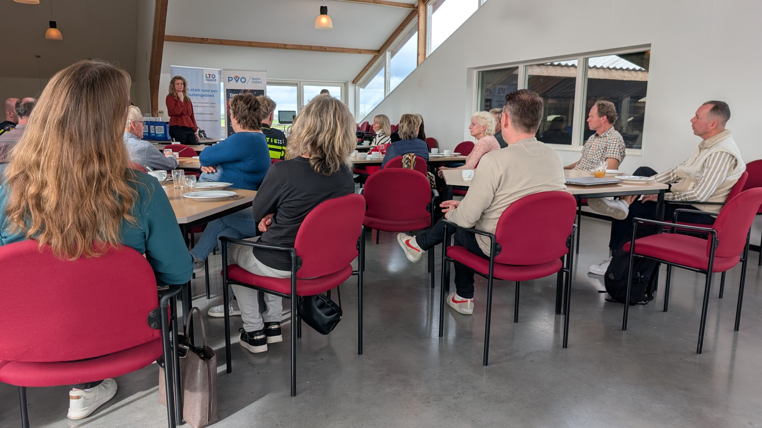 Mensen zitten op rode stoelen in een vergaderruimte en luisteren naar een vrouw die voor een groep spreekt. Aan de muur hangen banners met teksten. Tafels zijn gedekt met glazen, kopjes, en enkele borden.