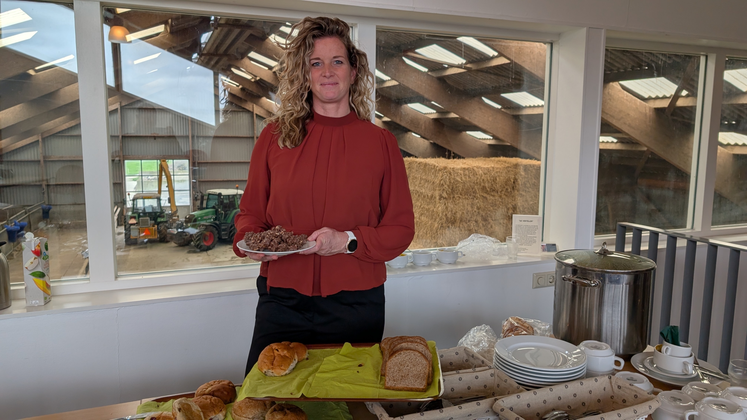 Een vrouw in een rode blouse houdt een bord met eten vast en staat voor een tafel met broodjes en servies, met op de achtergrond een loods met landbouwvoertuigen en hooibalen.