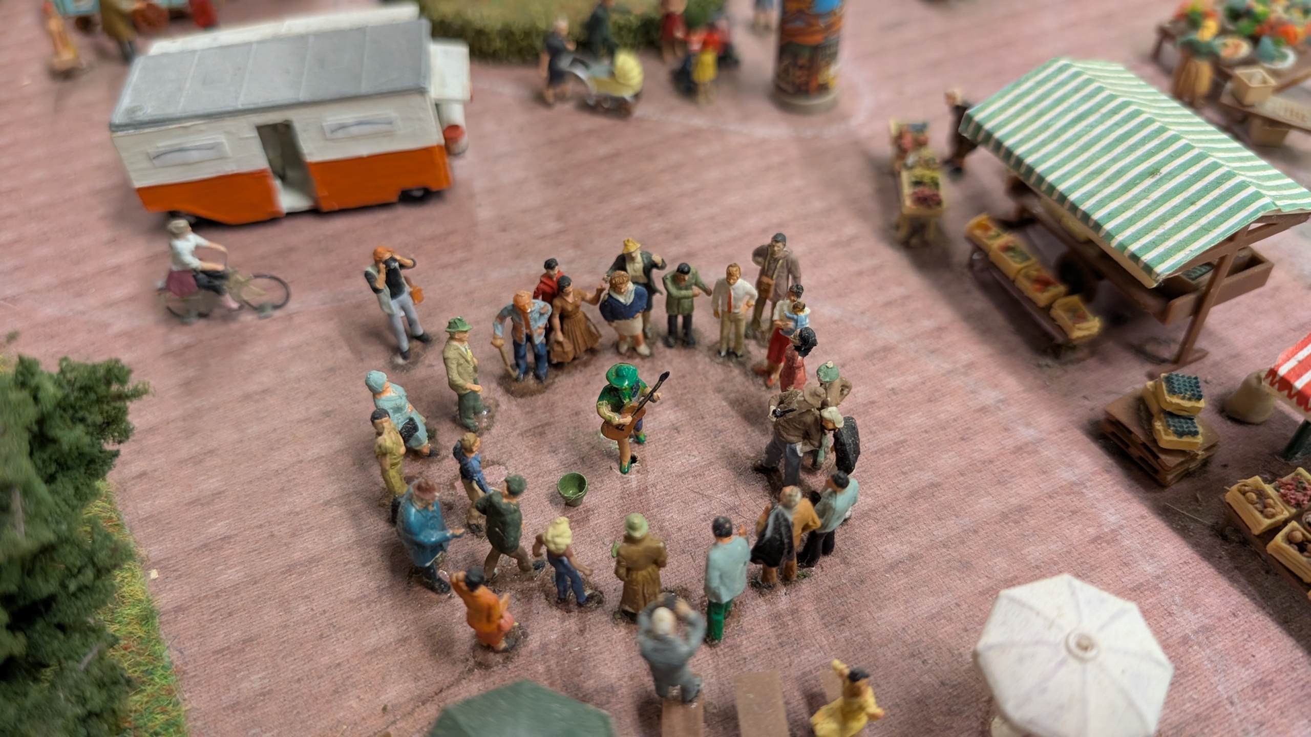 Miniatuur scène van een markt met een groep mensen die in een cirkel staan rond een muzikant met een gitaar. Er zijn kraampjes te zien met gestreepte overkappingen en een caravan op de achtergrond.