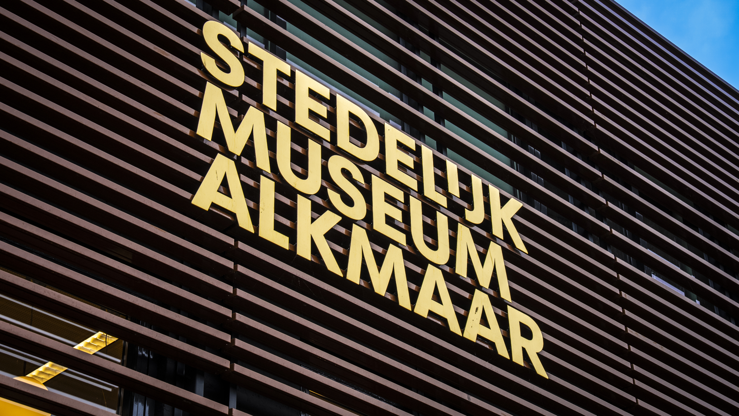 Gevel van het Stedelijk Museum Alkmaar met grote gele letters op een achtergrond van horizontale houten lamellen.