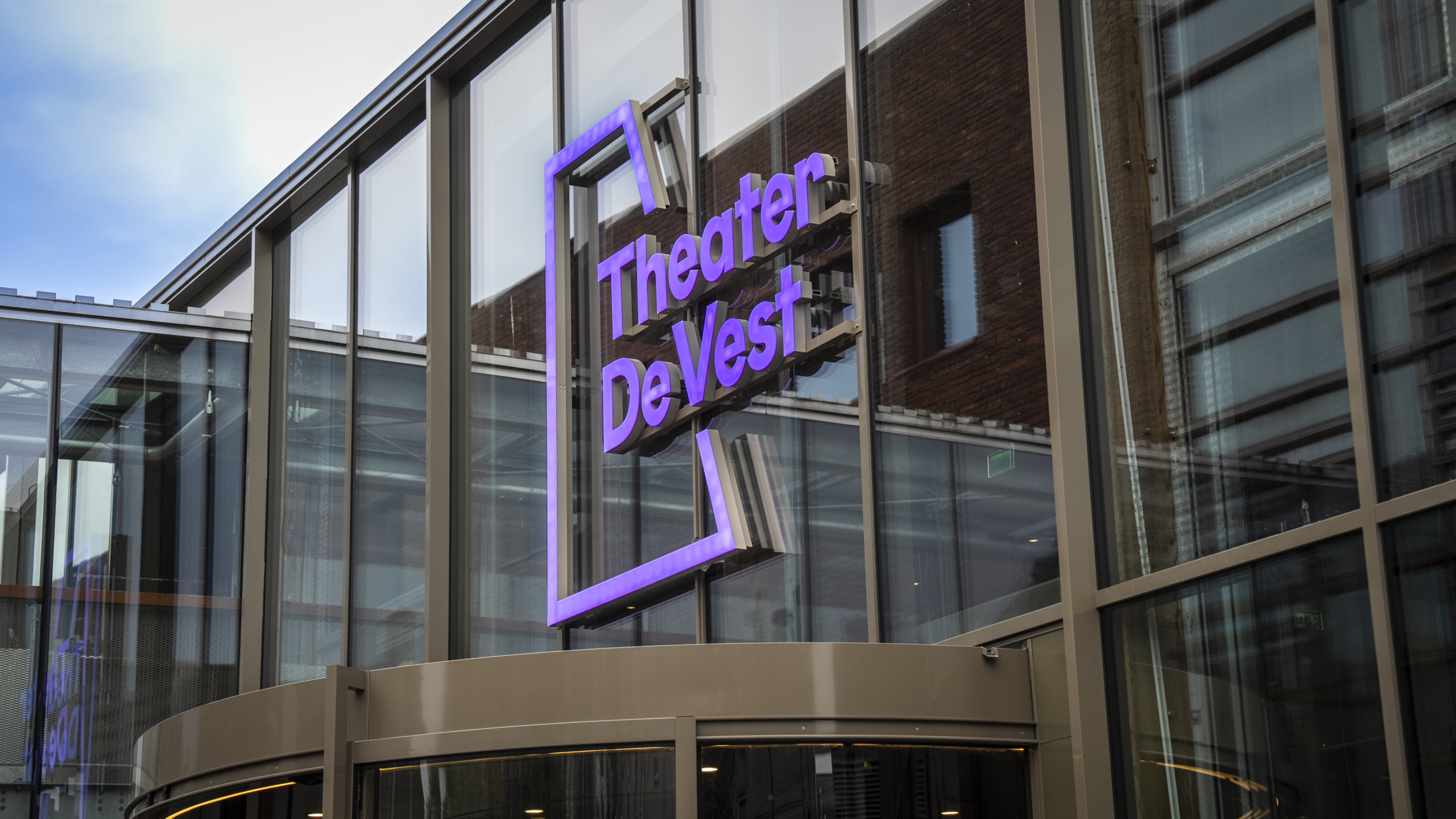 Buitenzijde van Theater De Vest met groot paars logo op glazen gevel.