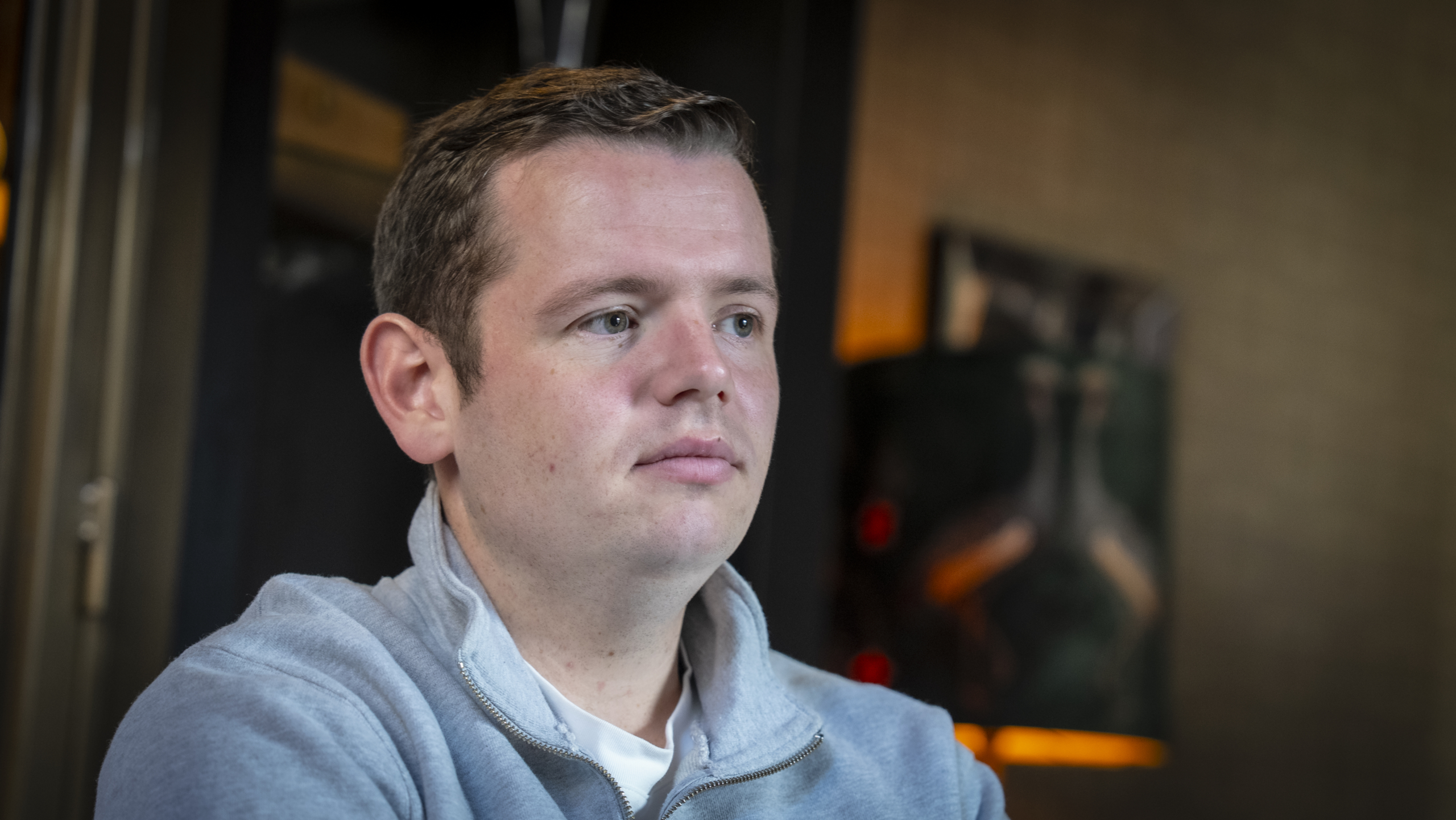 Man in a grijs vest kijkt naar links met een neutrale gezichtsuitdrukking, tegen een onscherpe achtergrond binnen.