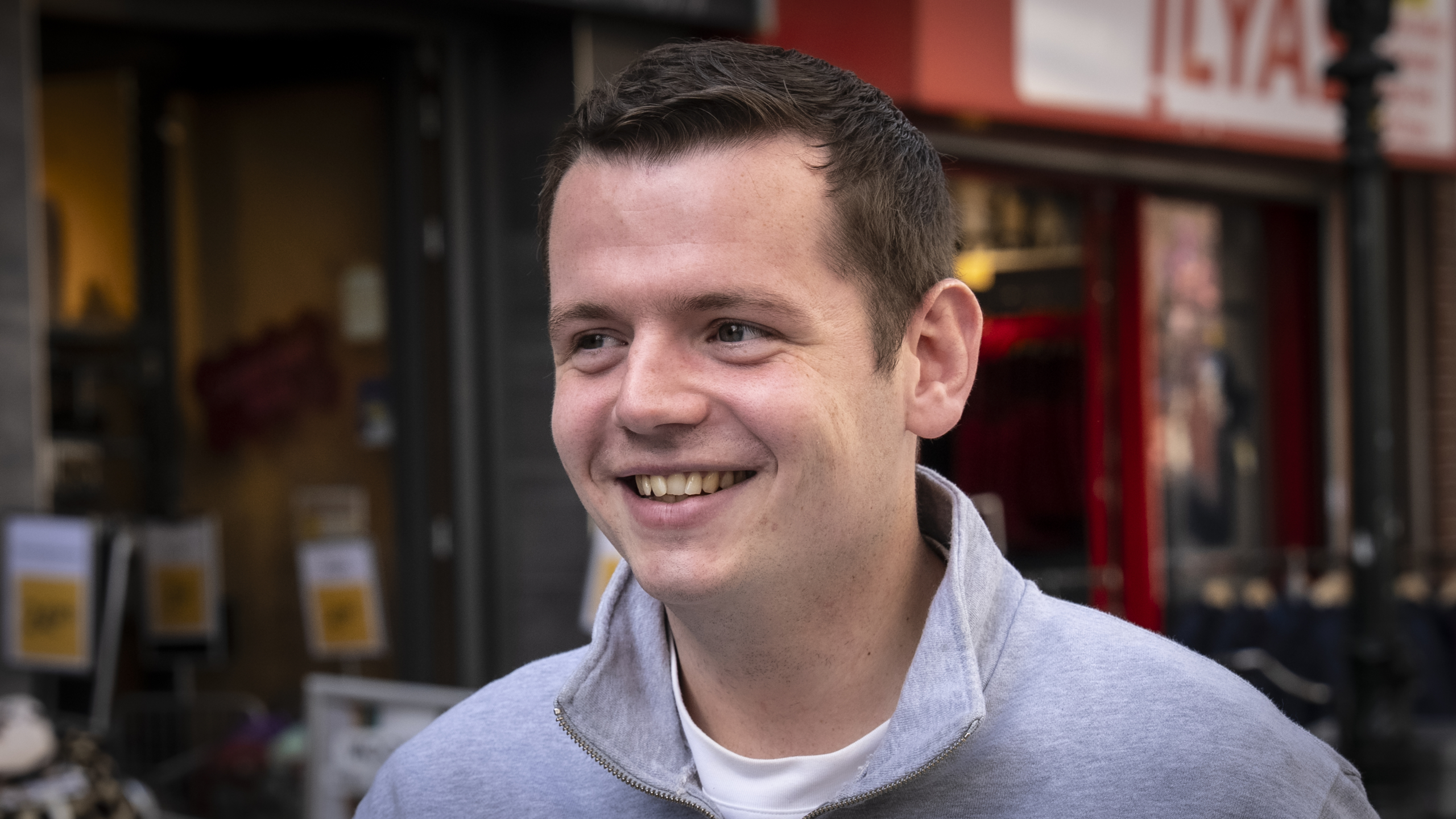 Jonge man met kort haar en grijs shirt lacht op straat voor een winkel met een felrode deur.