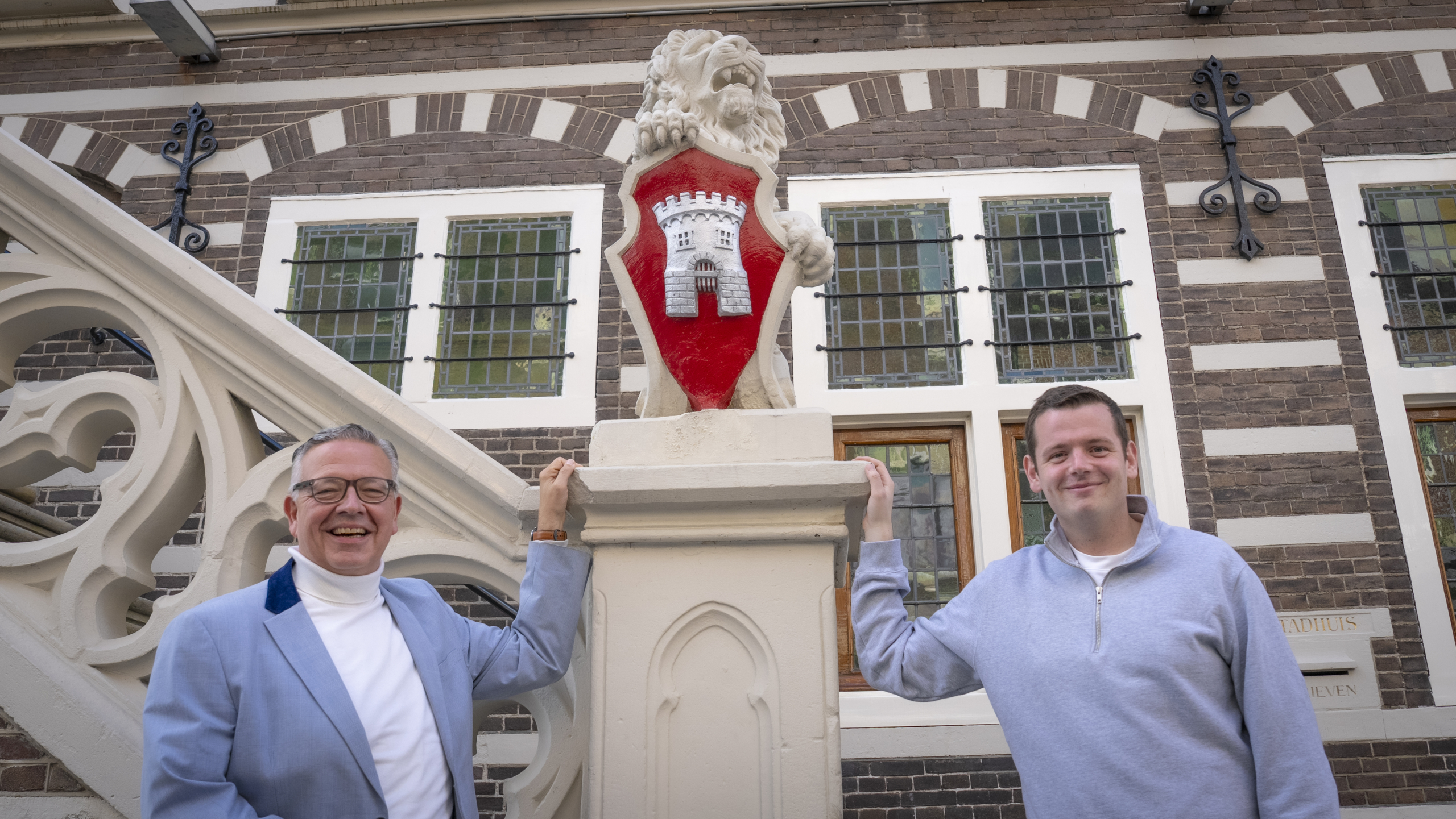 Twee mannen poseren lachend voor een decoratief beeld van een leeuw met een rood schild met een witte kasteelpoort, bij een historisch gebouw met bakstenen en glas-in-loodramen.