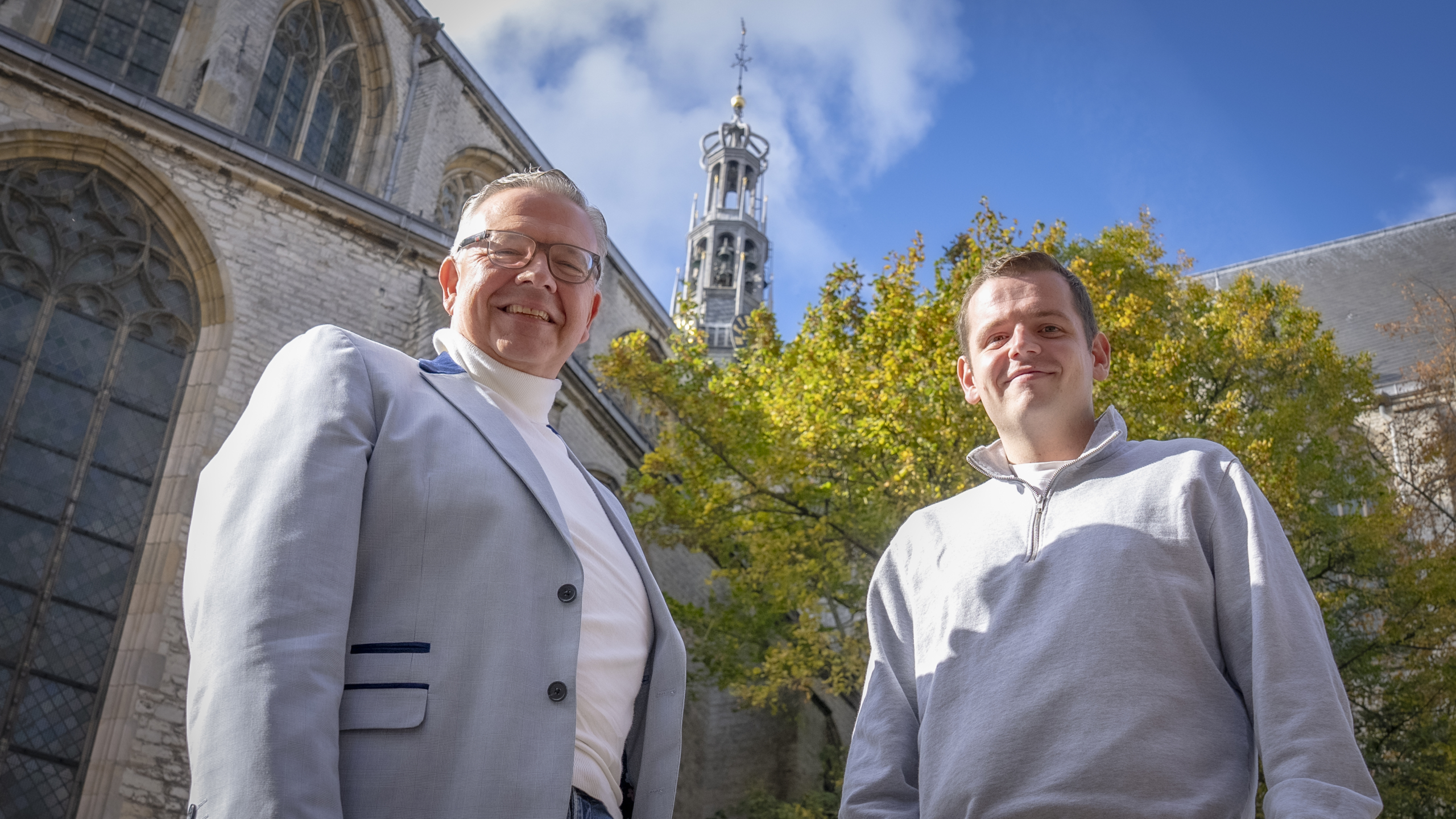 Twee mannen staan buiten voor een kerk of historisch gebouw, met een kloktoren en herfstbomen op de achtergrond. De man links draagt een lichtgrijs jasje en een coltrui, en de man rechts draagt een lichtgrijze trui. Ze lachen naar de camera onder een heldere blauwe hemel.