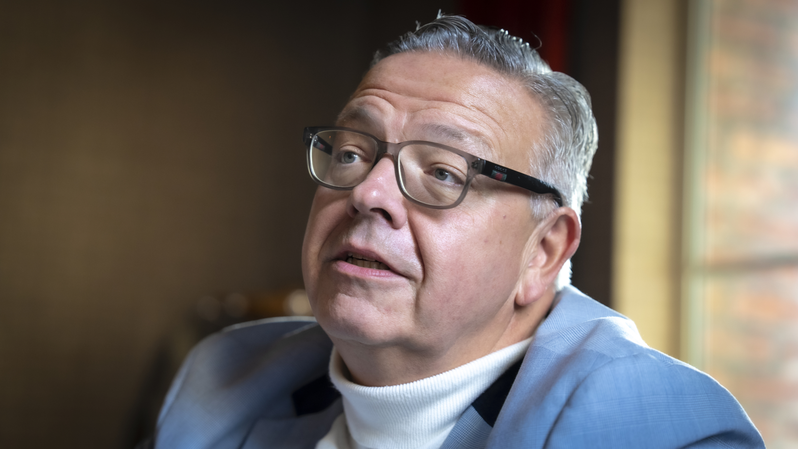 Een man met een bril en een grijs kapsel draagt een lichtblauw colbert en een witte coltrui, tegen een wazige achtergrond.