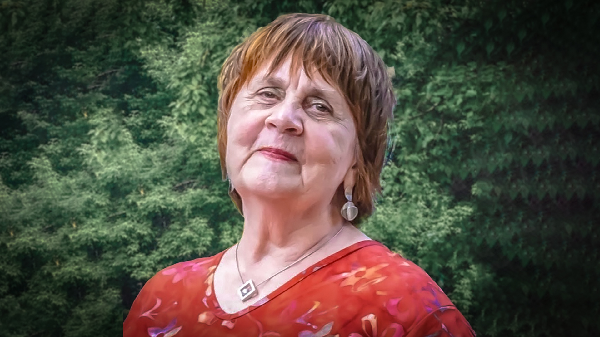 Oudere vrouw met kort, bruin haar en een rode blouse met bloemenmotief, staat buiten met een groene achtergrond van bomen.
