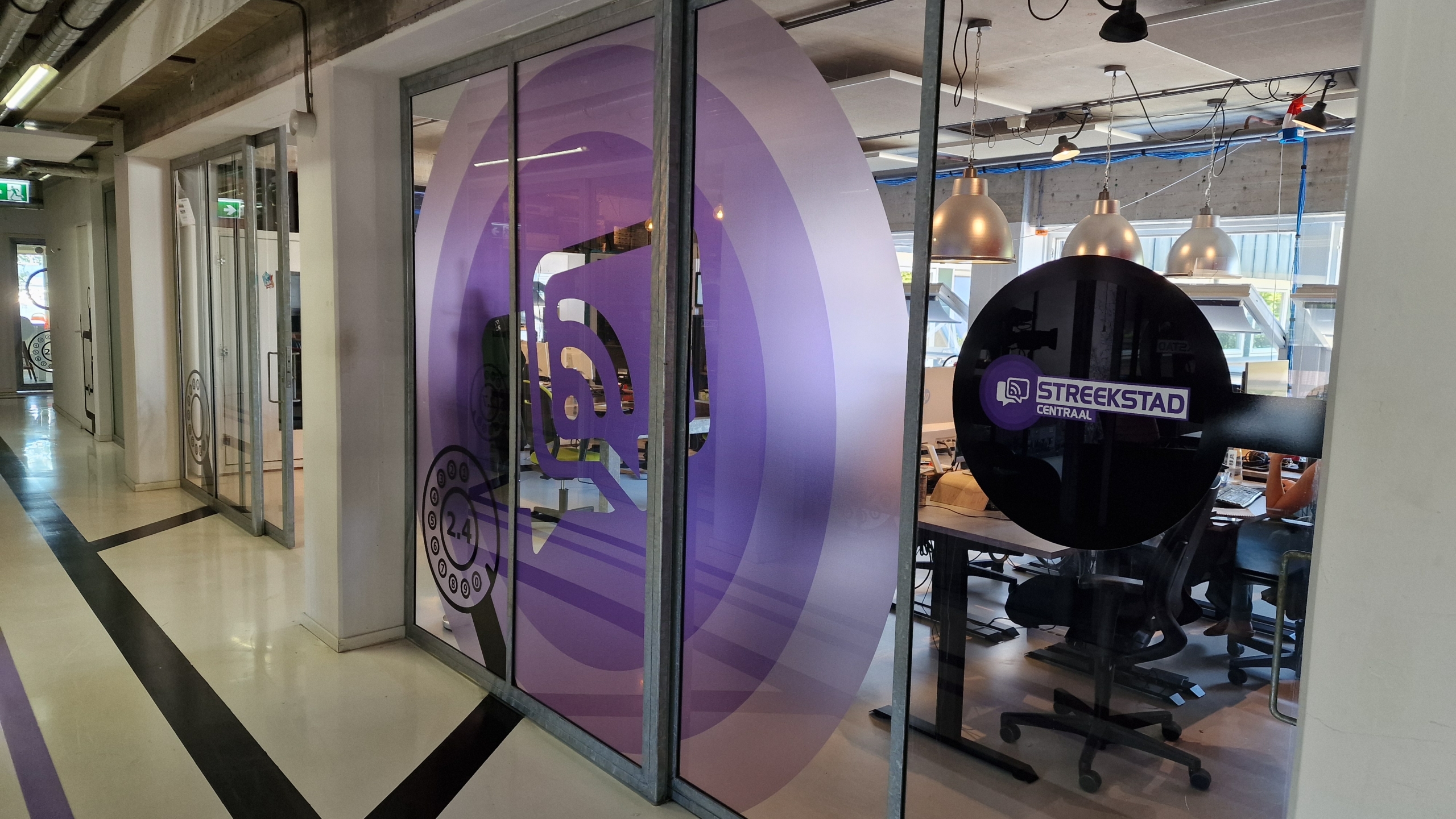 Een kantoorruimte met glazen wanden waarop paarse en zwarte ontwerpen zijn aangebracht, waaronder het logo "STREEKSTAD CENTRAAL". Binnen zijn bureaus, stoelen en computers zichtbaar, en aan het plafond hangen industriële lampen.
