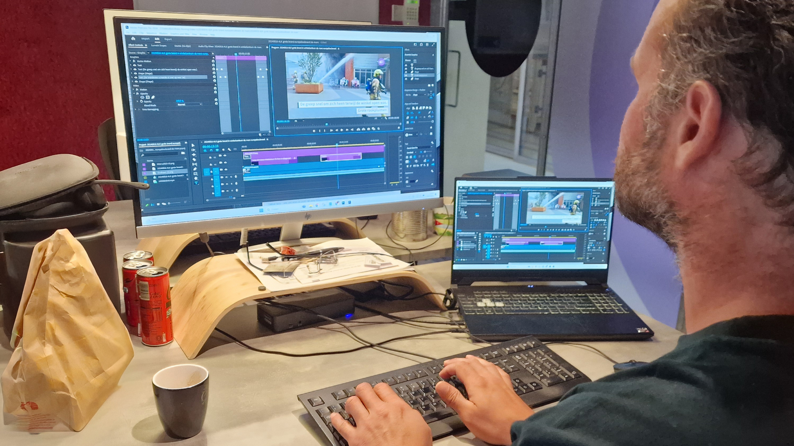 Een man aan een bureau werkt met twee schermen waarop videobewerkingssoftware is geopend, omgeven door koffie, cola en papierwaren.