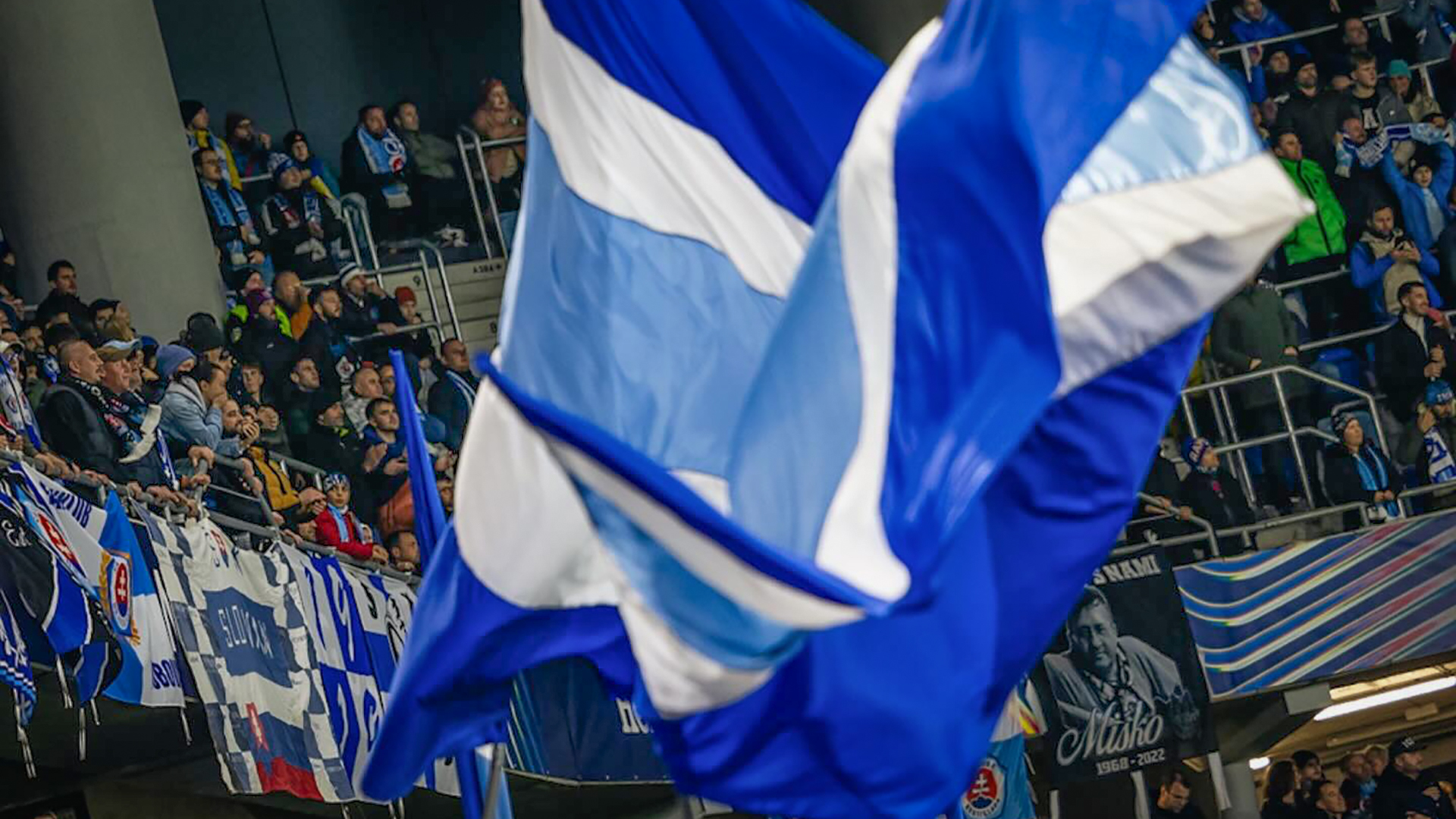 Een menigte van enthousiaste voetbalsupporters in een stadion, met blauwe en witte vlaggen die wapperen en Duitse vlaggen op de achtergrond.