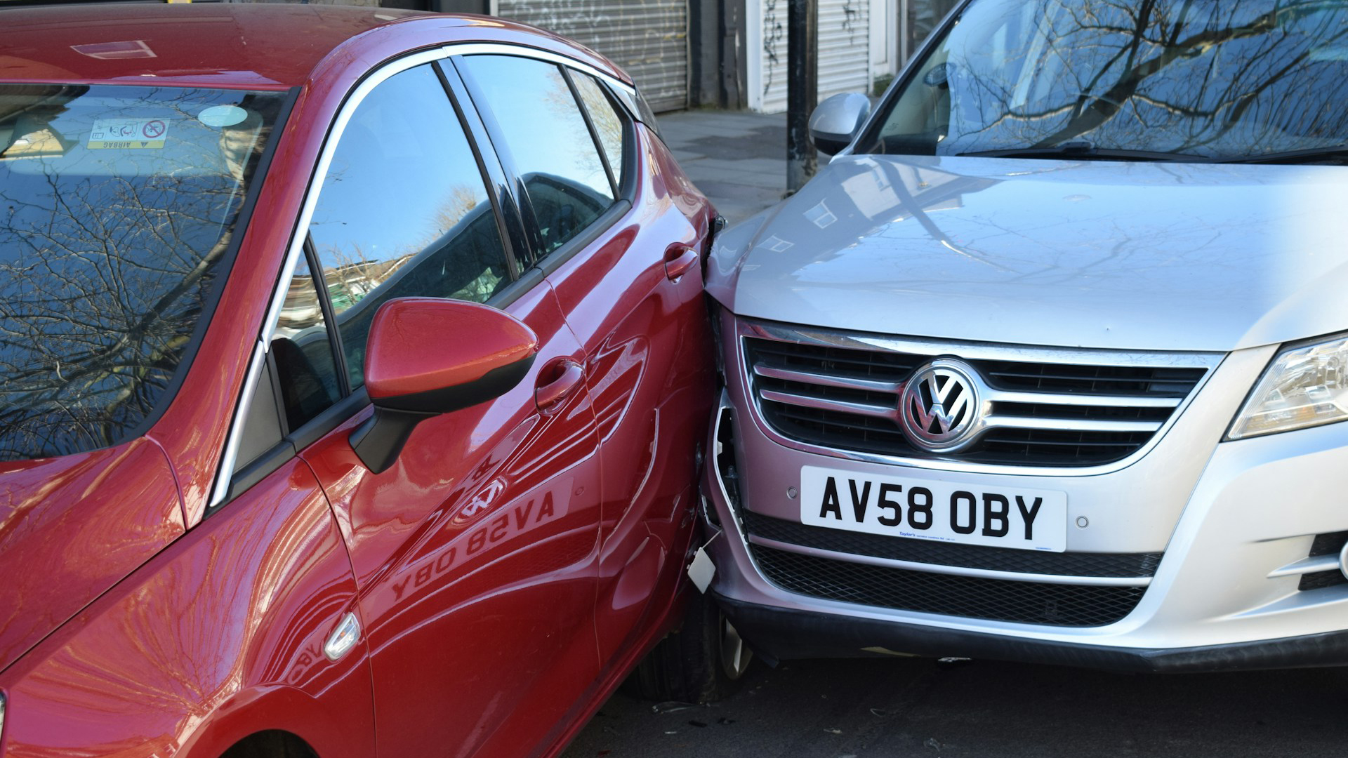 Een rode auto en een zilverkleurige auto met een Volkswagen-embleem zijn met elkaar in botsing gekomen, waarbij de zijkant van de rode auto is beschadigd.
