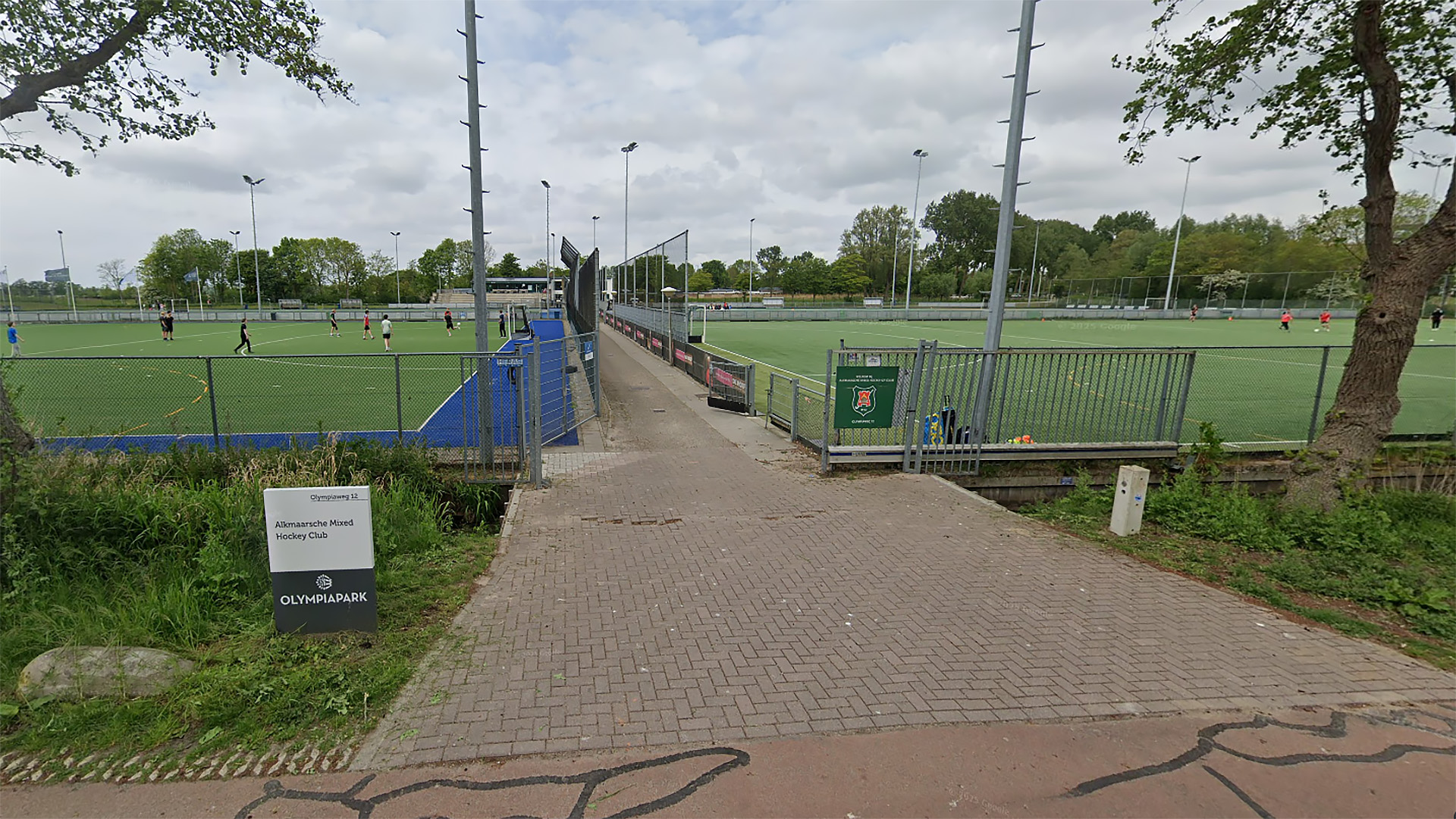 Ingang naar een hockeyveld met enkele hockeyspelers in beweging, naast een bord met de tekst 'Alkmaarsche Mixed Hockey Club' en 'Olympiapark'. Het veld is omgeven door een hek en vlaggenmast, met bomen en een bewolkte lucht op de achtergrond.