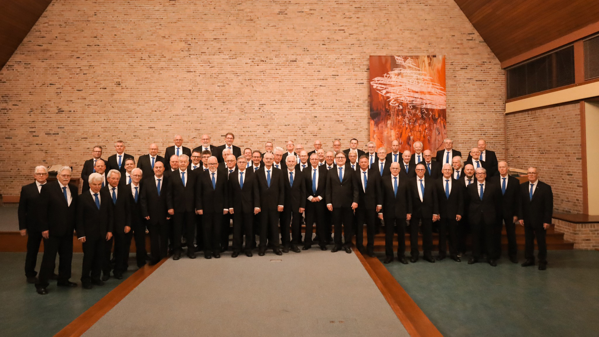 Een grote groep mannen in zwarte pakken met blauwe dassen poseren samen in een kerkelijk gebouw met een bakstenen muur en een abstract schilderij op de achtergrond.