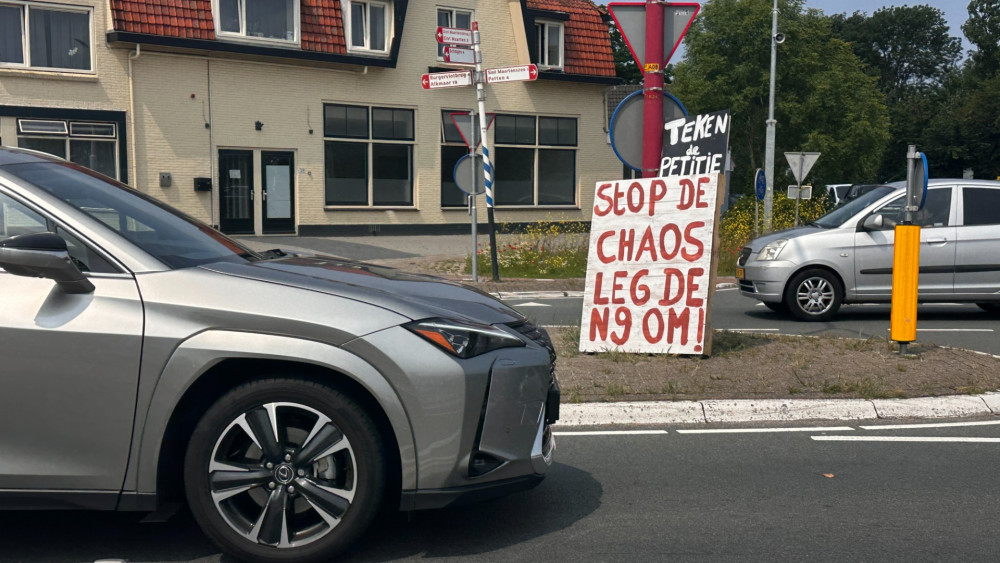 Een verkeersbord midden op een rotonde toont de tekst 'Stop de chaos leg de N9 om!' in rode letters. Op de achtergrond zijn een gebouw en meerdere voertuigen zichtbaar.