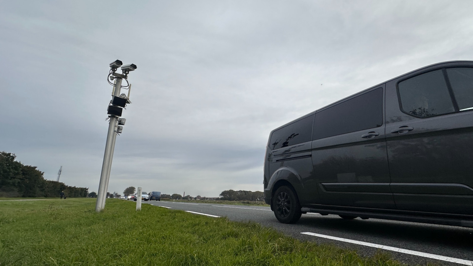 Een donkere bestelwagen rijdt op een landelijke weg, naast een paal met meerdere camera's erop gemonteerd, met een bewolkte lucht op de achtergrond.
