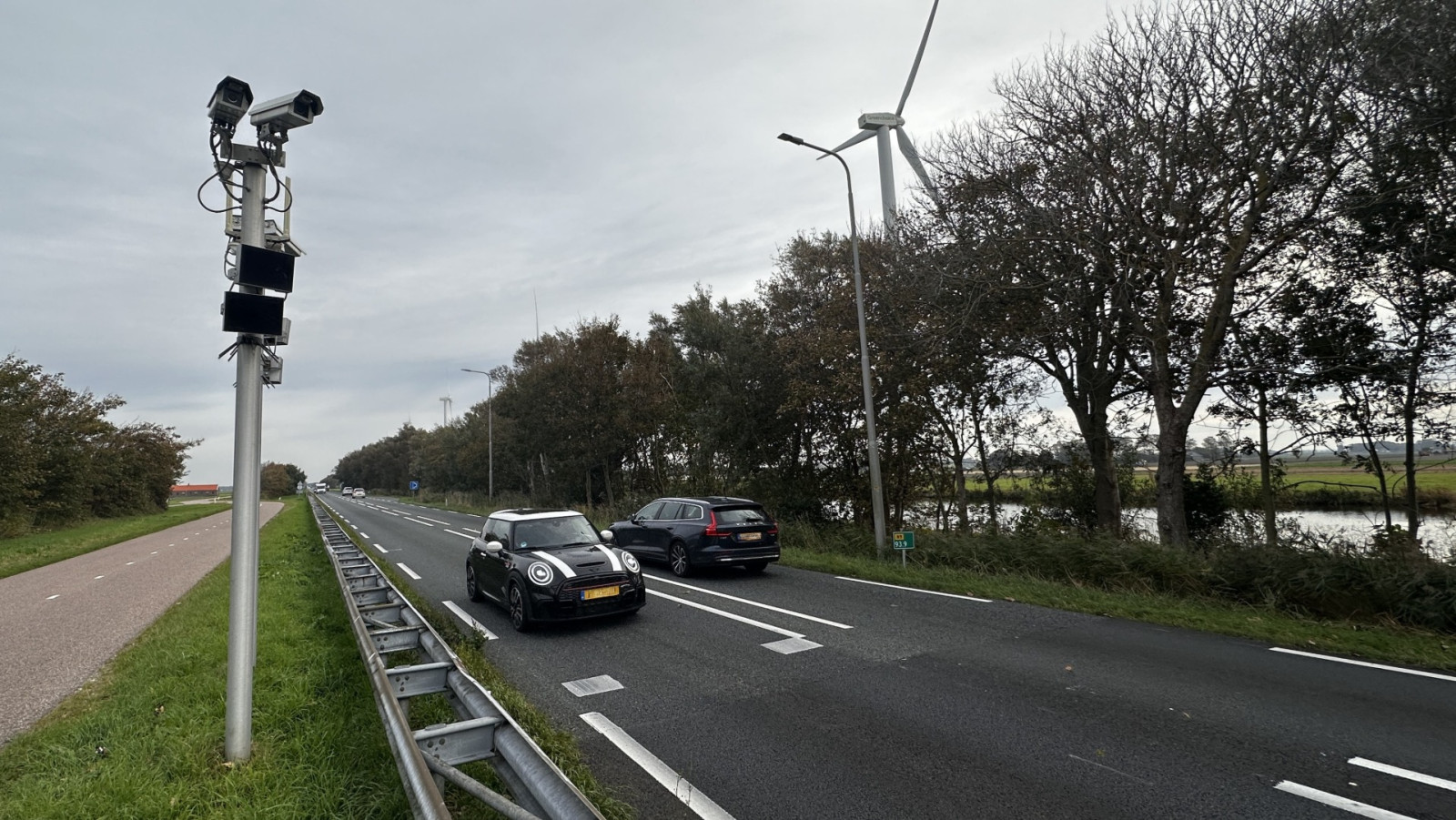 Twee auto's rijden op een tweebaansweg langs een rij bomen, met een beveiligingscamera op een paal aan de linkerkant van het beeld.
