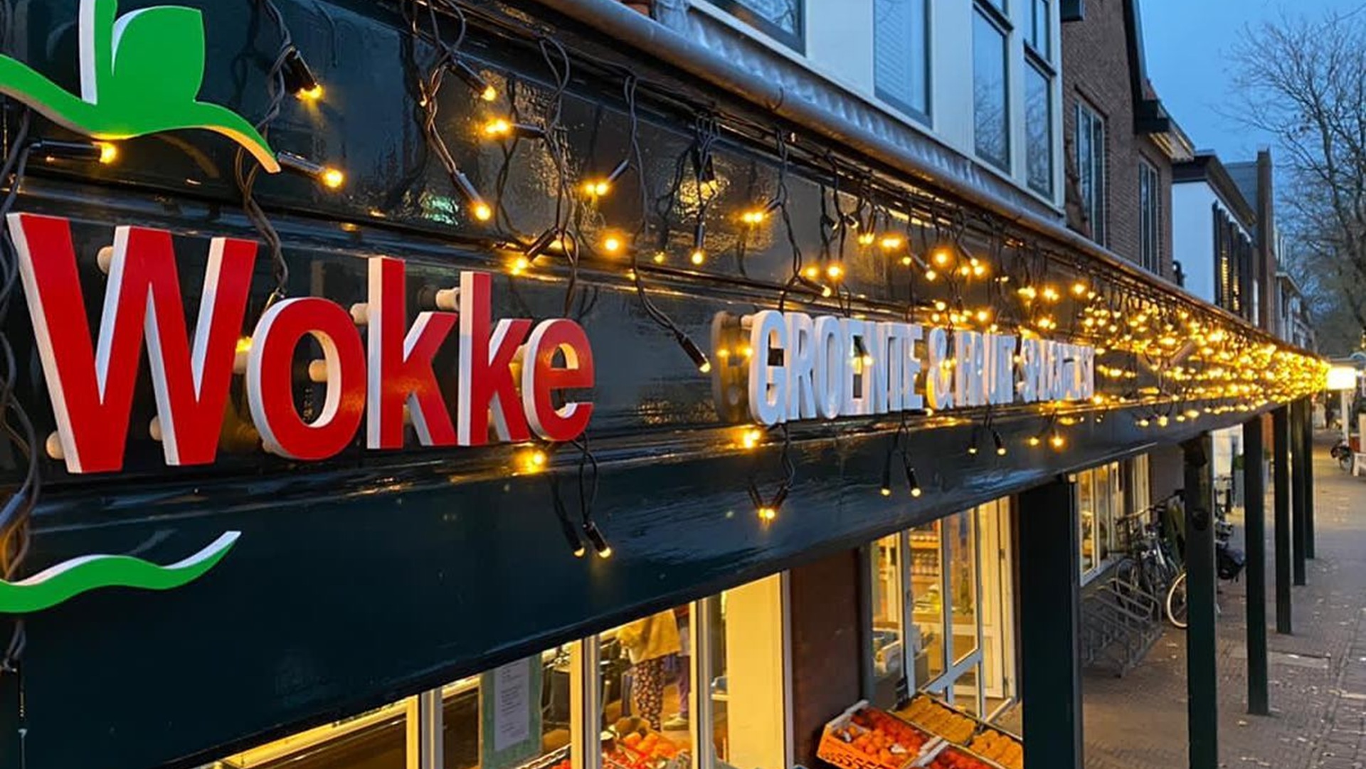 Verlicht winkeletalage van Wokke, een groente- en fruitwinkel, met kerstverlichting boven de etalage en fruitkisten buiten op de stoep.