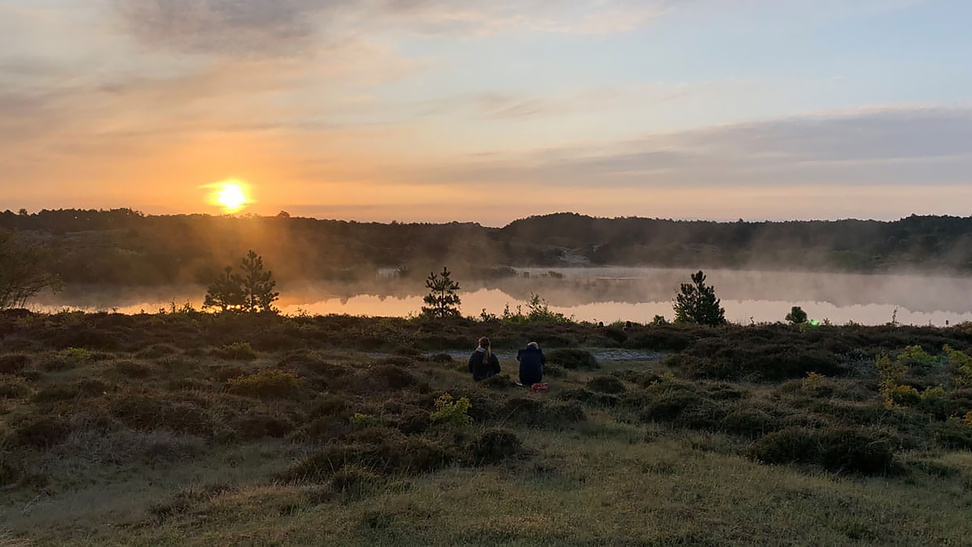 Twee mensen zitten op een grasgebied bij zonsopgang, met zicht op een mistige vijver en bomen in de verte.