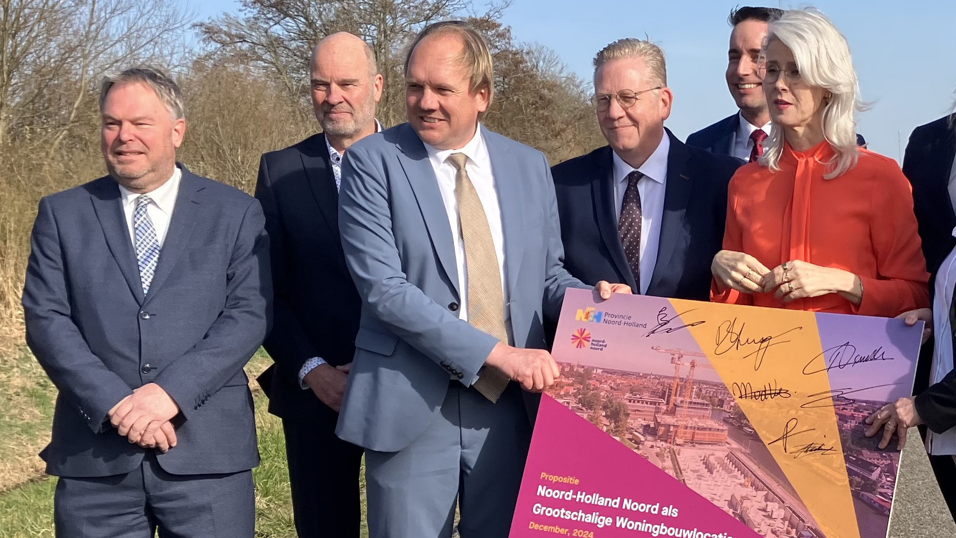Groep mensen in formele kleding houdt een poster vast met de tekst over een voorstel voor grootschalige woningbouwlocaties in Noord-Holland Noord, gedateerd december 2024, ondertekend door diverse personen.