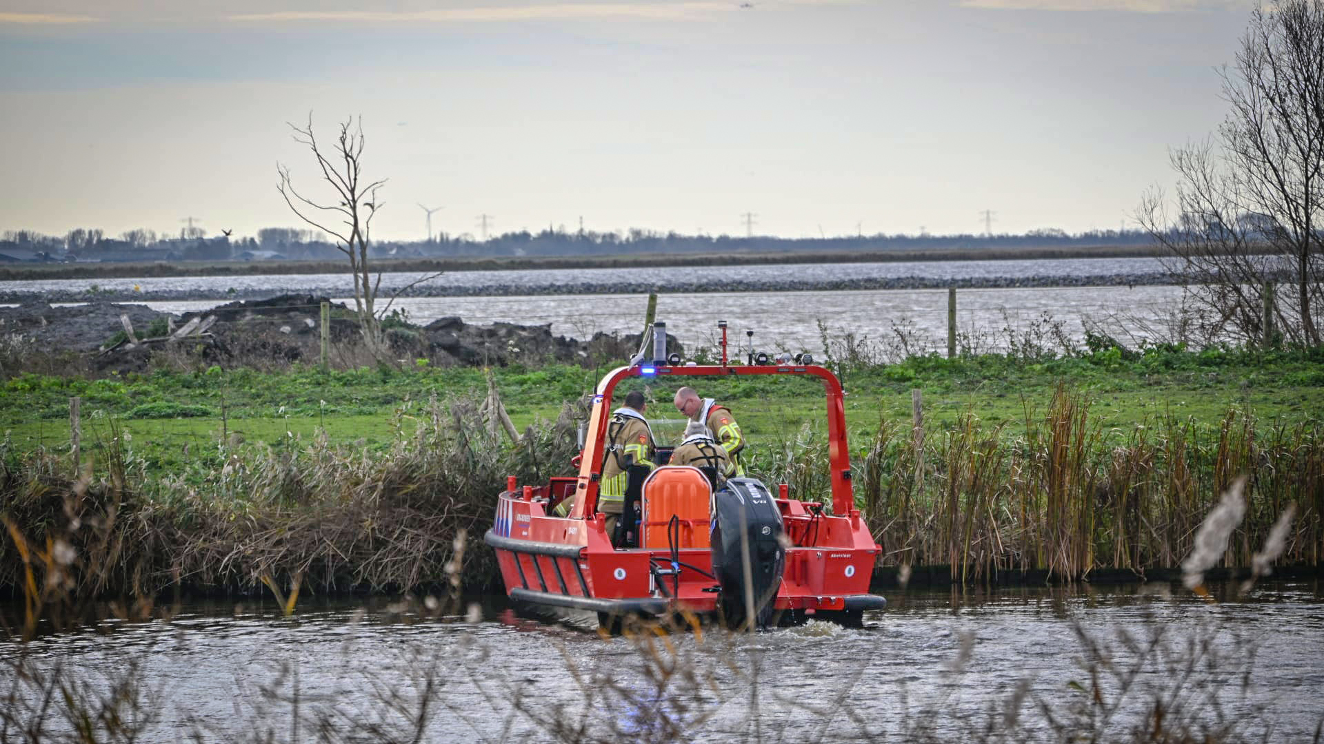 Twee mensen in reddingskleding staan op een kleine rode reddingsboot op een rivier, met een weids landschap en een kale boom op de achtergrond.