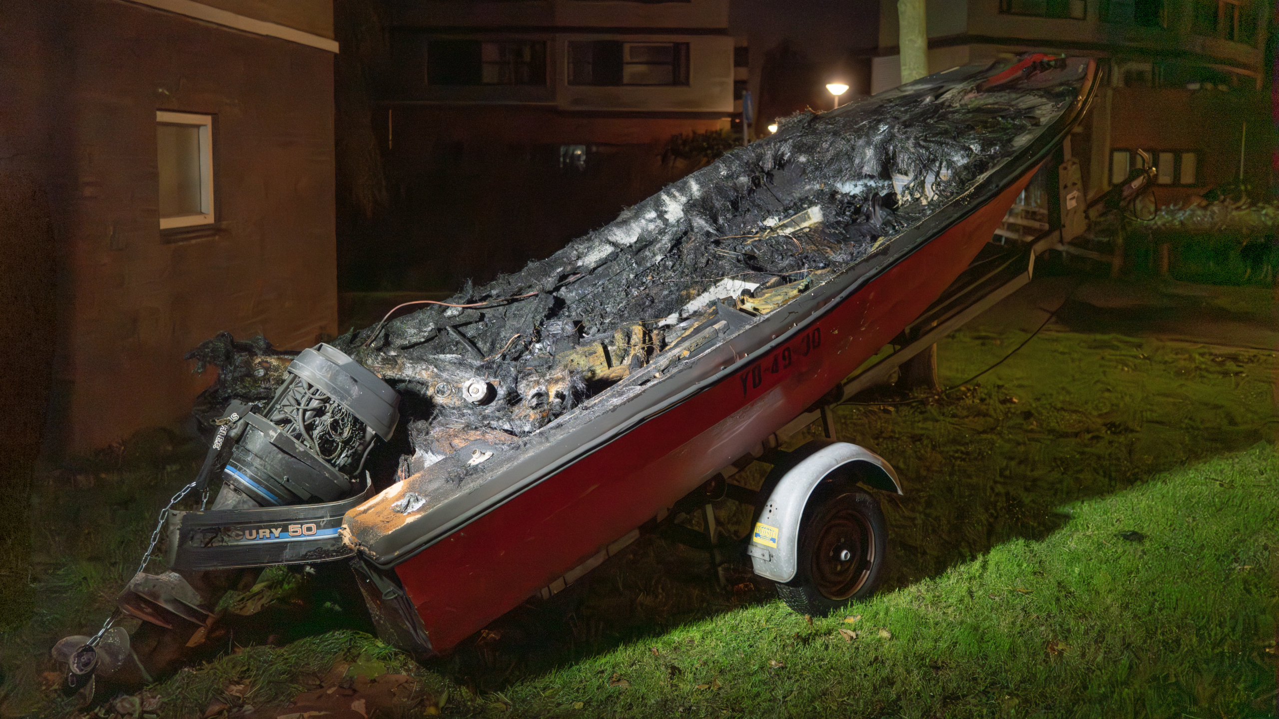Sterk beschadigde boot met verbrande binnenkant geparkeerd in een residentiële omgeving 's nachts, met een buitenboordmotor zichtbaar aan de achterkant.