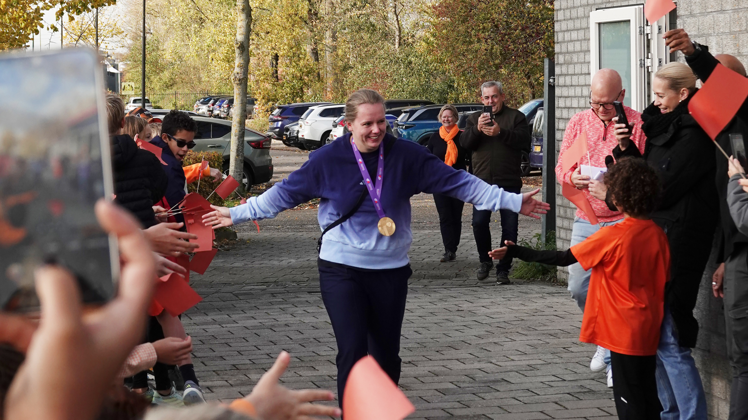 Vrouw met medaille loopt lachend door een rij mensen buiten, waarbij ze handen schudt terwijl ze wordt toegejuicht.