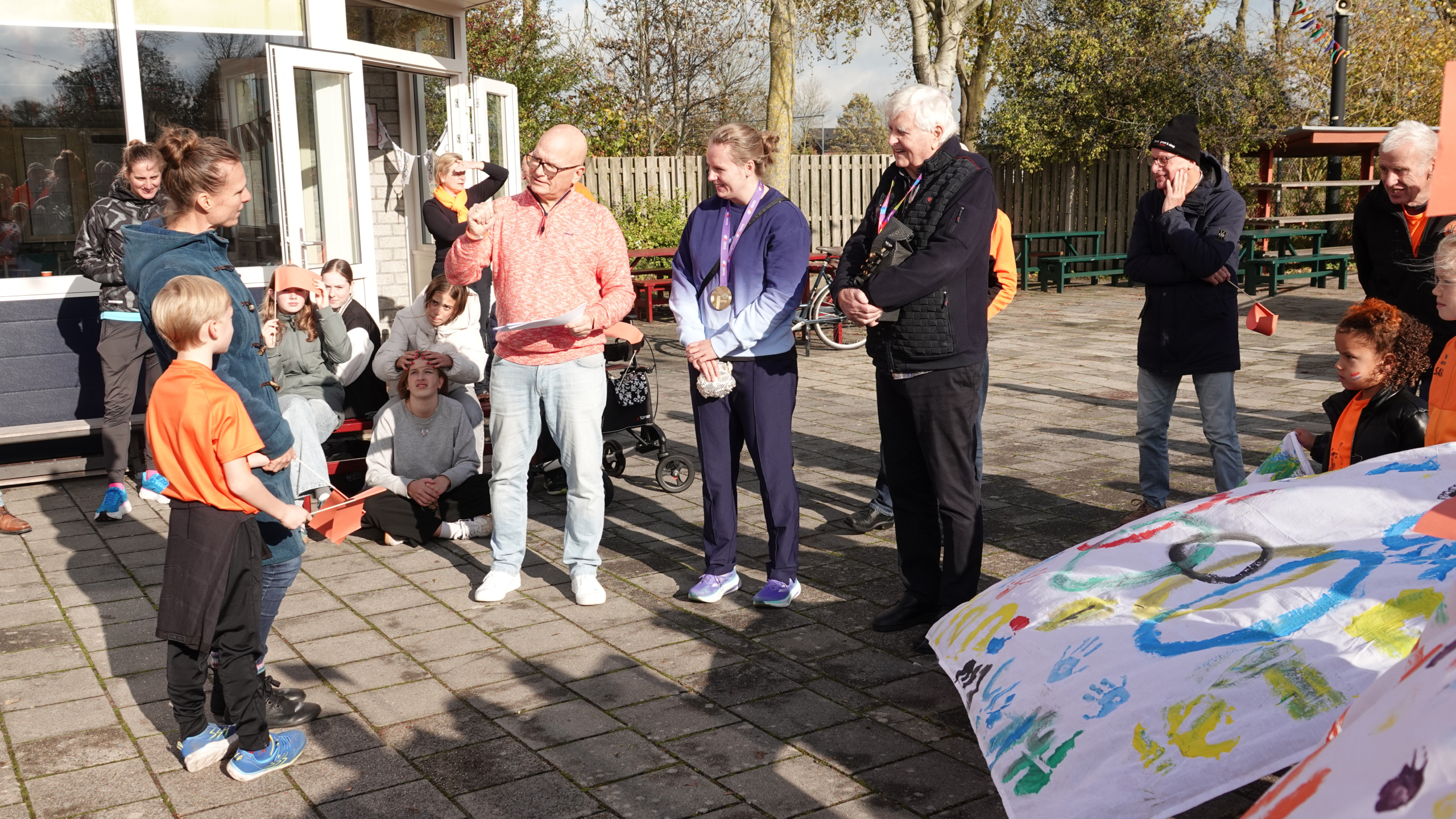 Een groep mensen, waaronder een vrouw met een medaille om haar nek, verzameld buiten bij een schoolgebouw. Een jong kind in een oranje shirt en een vrouw in een blauwe jas luisteren naar een man die iets voorleest. Andere mensen zitten op een bank en kijken toe, met een kleurrijk geschilderd doek in de voorgrond.