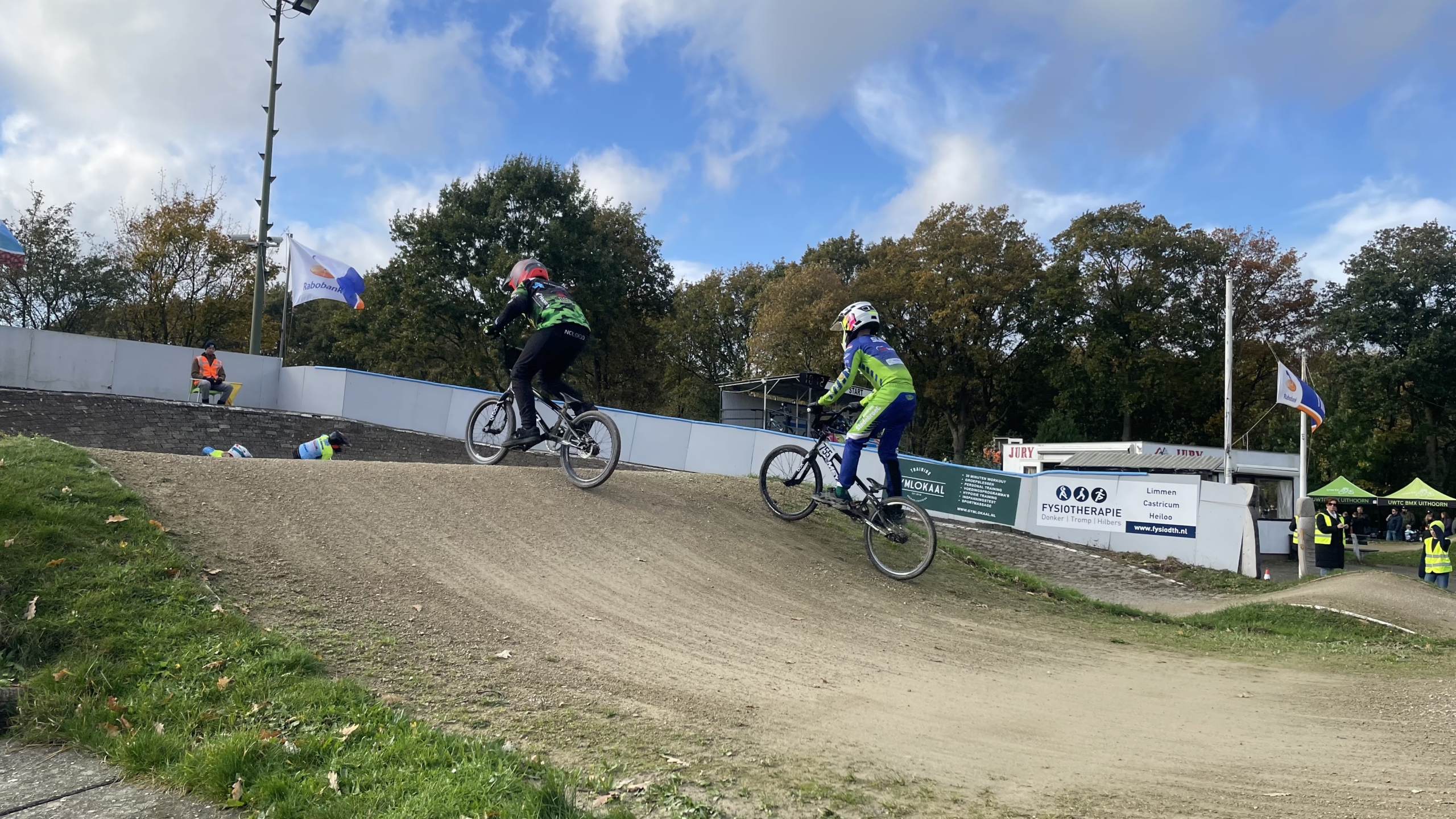 Twee BMX-rijders in actie op een heuvelachtige baan, omgeven door bomen en enkele toeschouwers in gele hesjes. Er zijn vlaggen en borden zichtbaar langs het parcours.