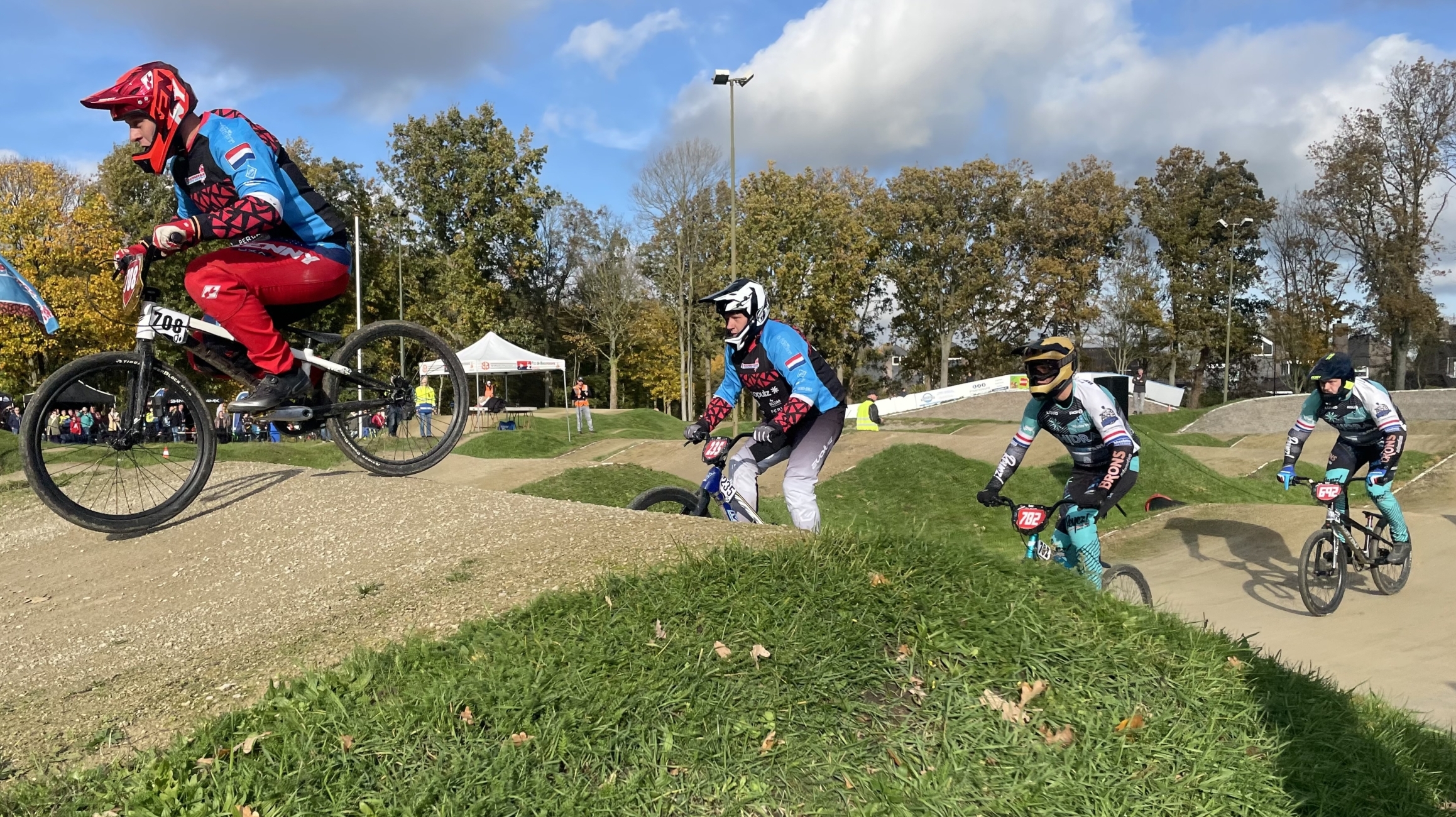BMX-fietsers in felgekleurde pakken racen over een heuvelachtig parcours met grasrijke delen en een heldere blauwe hemel op de achtergrond.