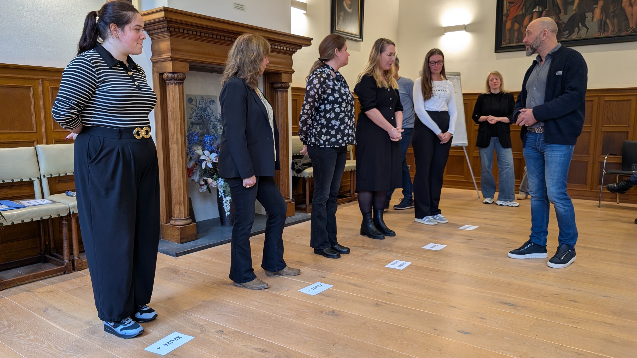 Groep mensen staat in een kamer met houten vloer en een haard, kijkend naar een man die voor een instructeur lijkt te staan. Op de vloer liggen papieren met woorden erop.