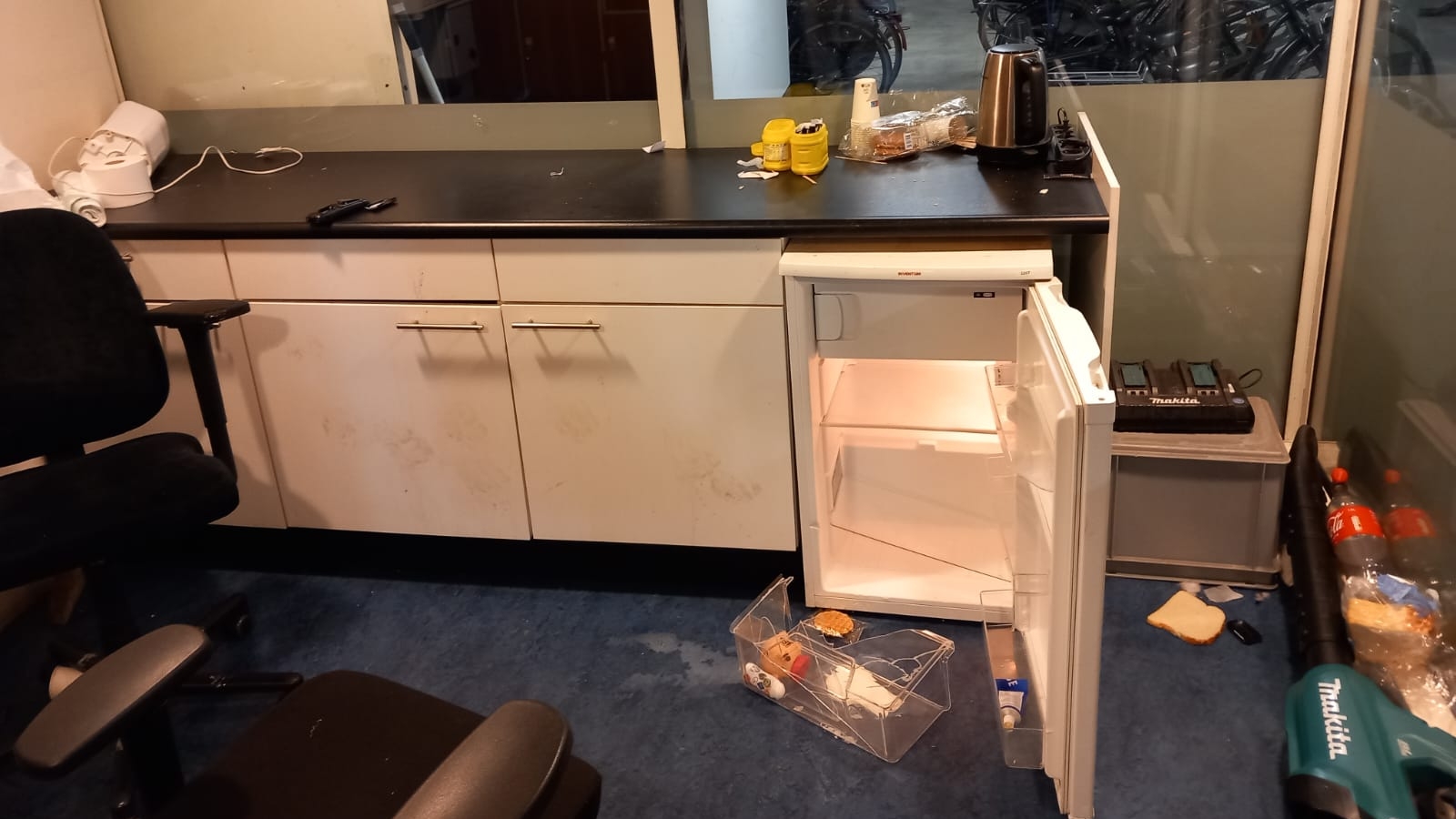 Een kantoorruimte met een bureau en opbergkast met open koelkast, waar enkele voedingsmiddelen, een waterkoker en een Makita gereedschapskoffer op staan; door de glazen wand is een fietsenstalling te zien.