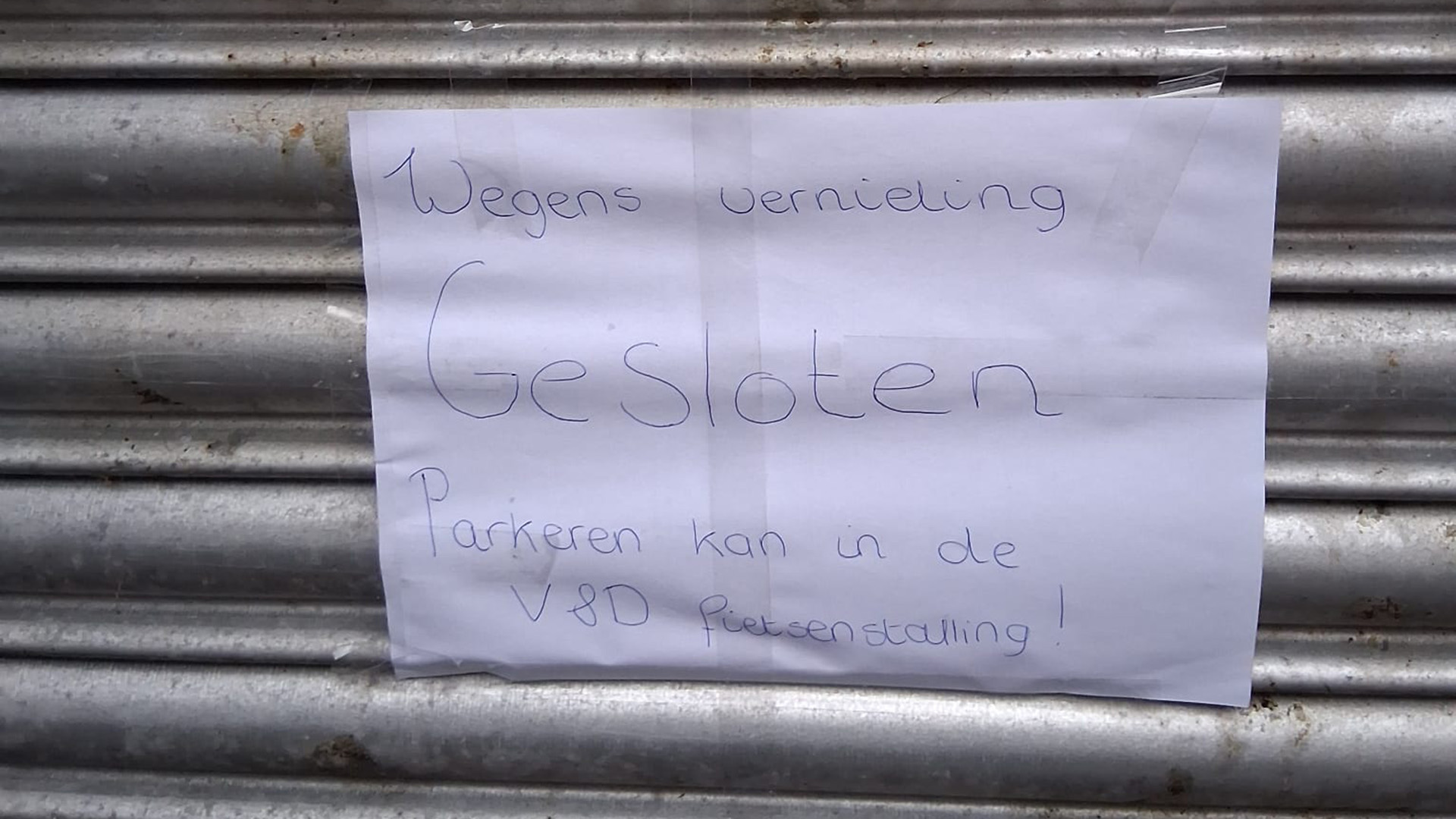 Een handgeschreven briefje op een metalen garagedeur met de tekst: 'Wegens vernieling gesloten. Parkeren kan in de V&D fietsenstalling!'