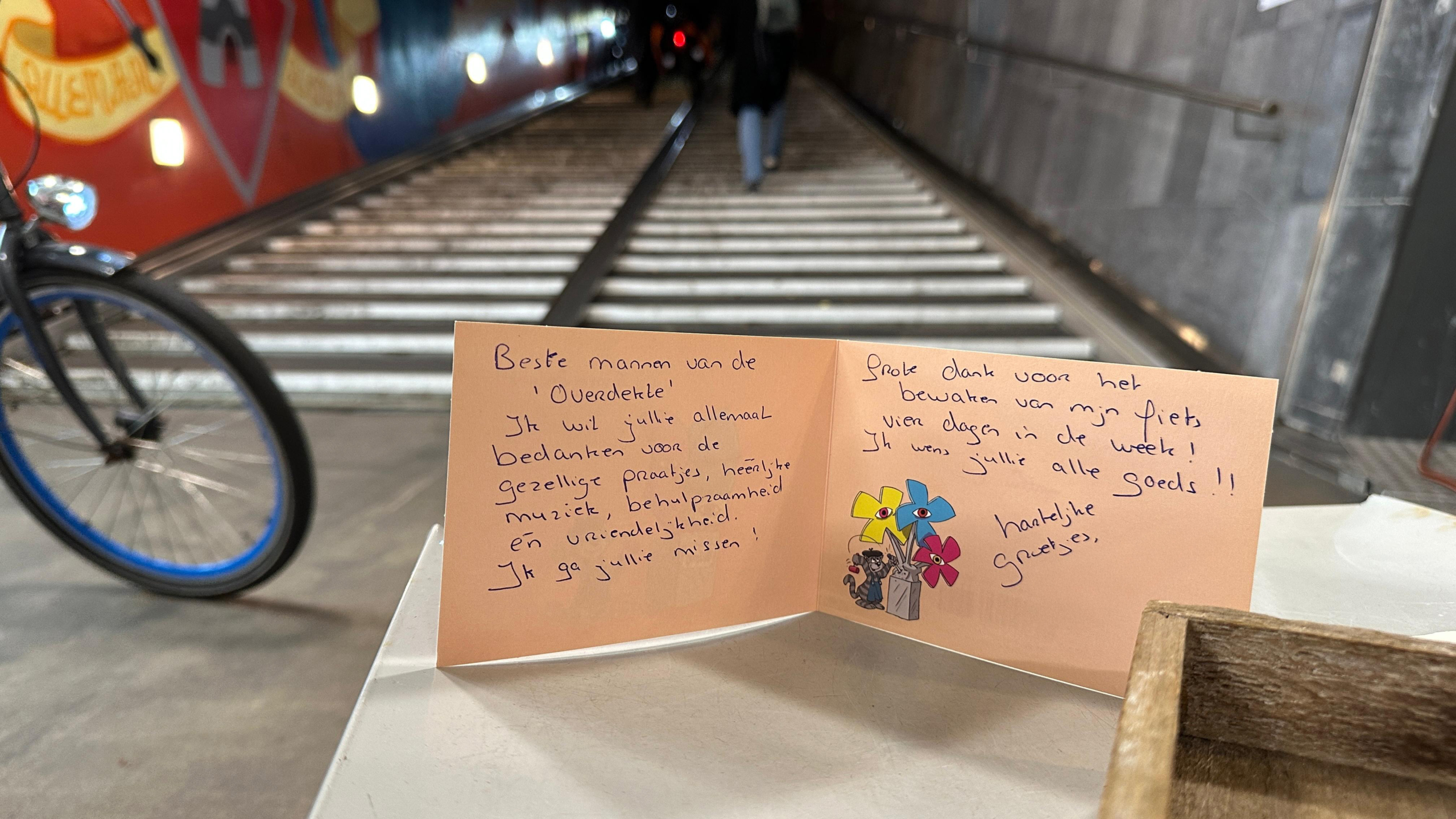 Een bedankkaart met handgeschreven tekst staat op een tafel naast een fietstunnel met een felgekleurde muurschildering en een fiets op de achtergrond. Op de kaart staan berichten van dankbaarheid voor vriendelijkheid en hulp.