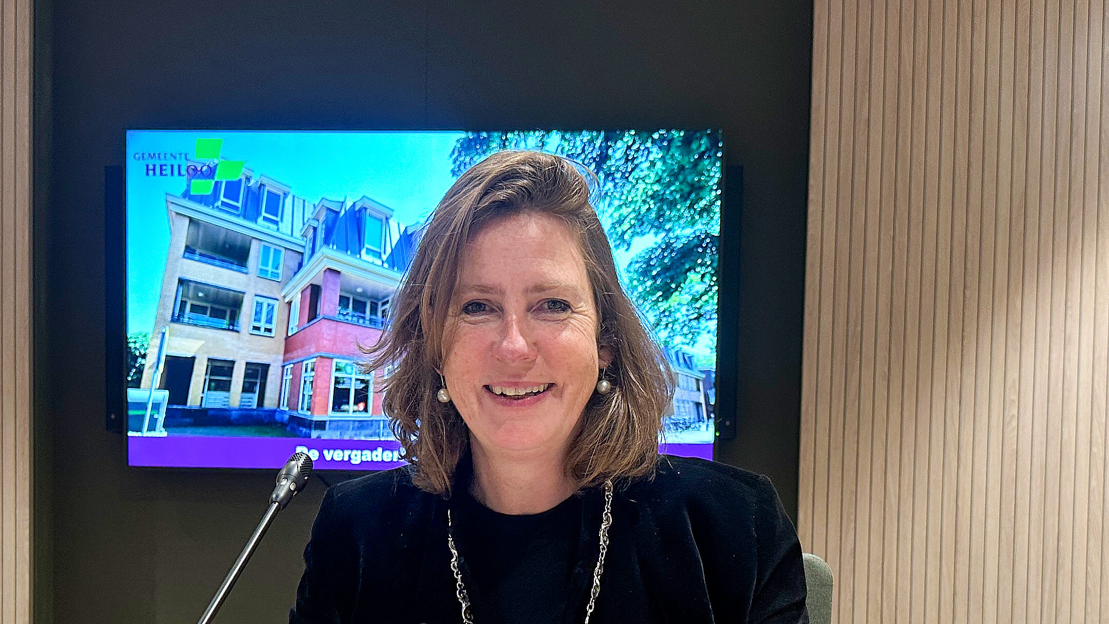 Vrouw glimlachend voor een microfoon met een scherm op de achtergrond waarop een gebouw en de tekst 'Gemeente Heiloo' te zien zijn.