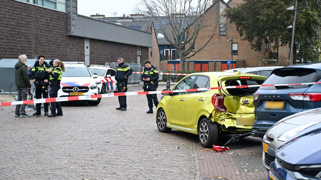 Politieagenten staan bij een afgezette plaats van een ongeval, met een beschadigde gele auto en politievoertuigen op de achtergrond.