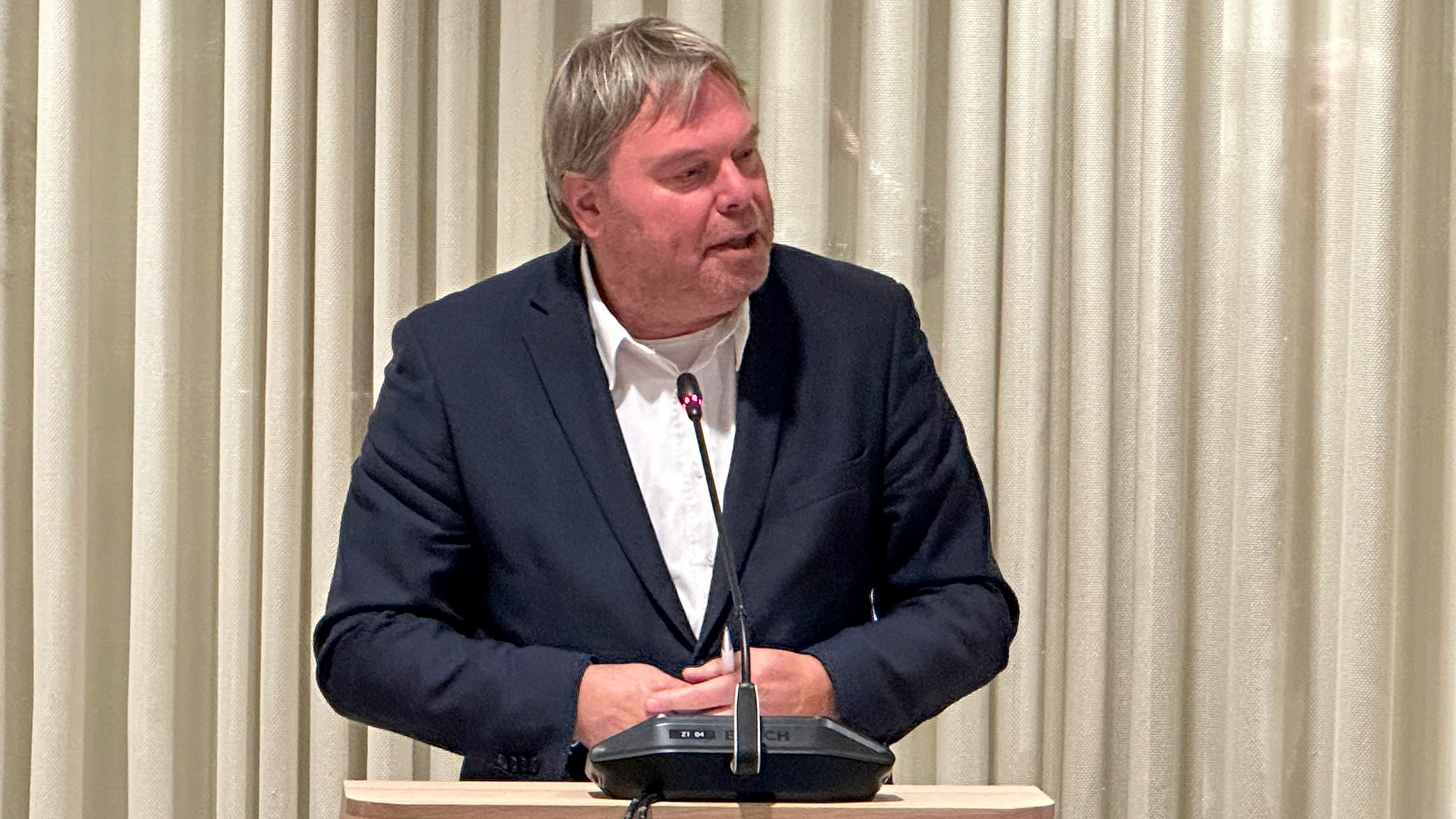 Een man in een donker pak spreekt bij een lessenaar met een microfoon, voor een achtergrond van lichte gordijnen.