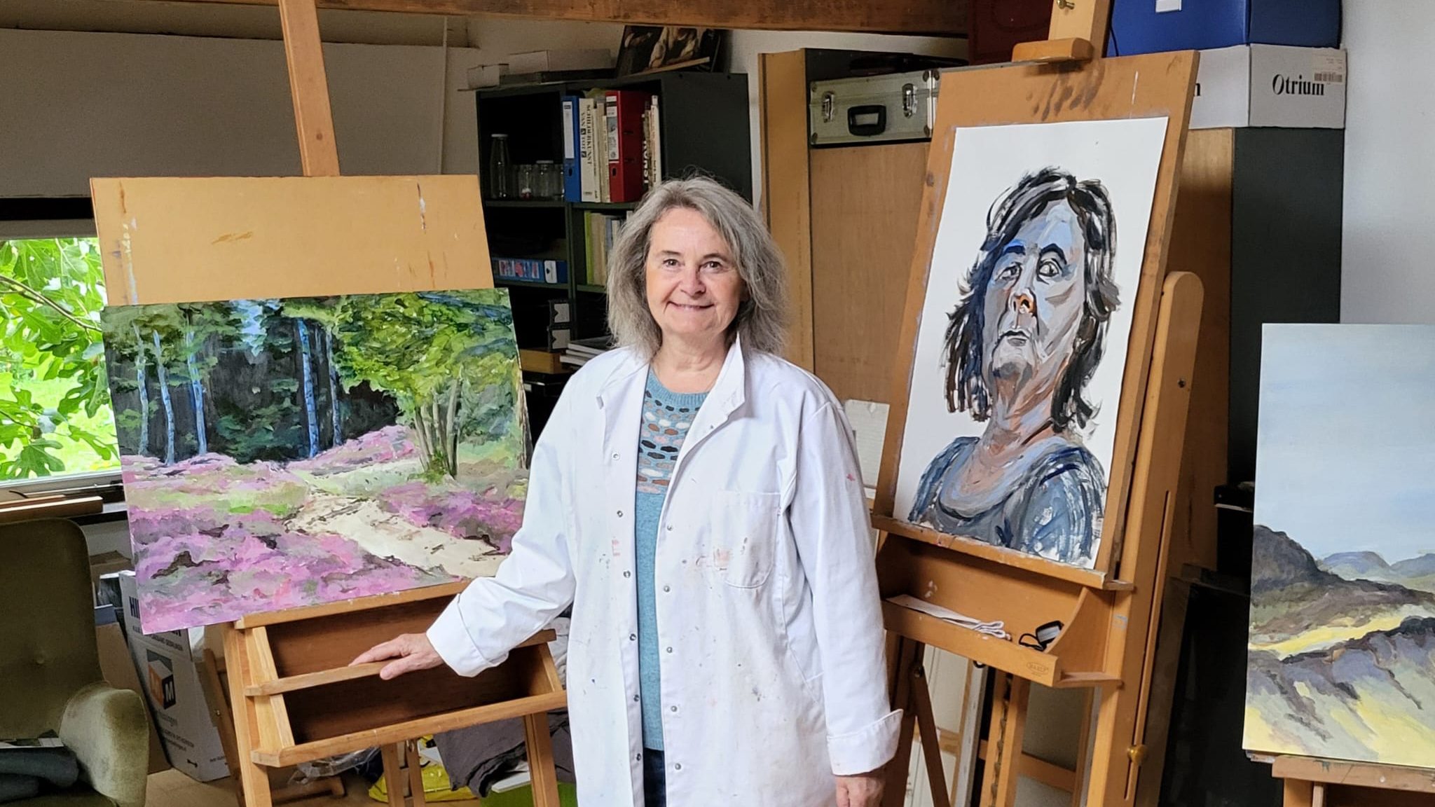Een vrouw in een wit schildersjas staat in een atelier tussen schildersezels met schilderijen; links een landschap met bomen en bloemen, rechts een expressief portret.