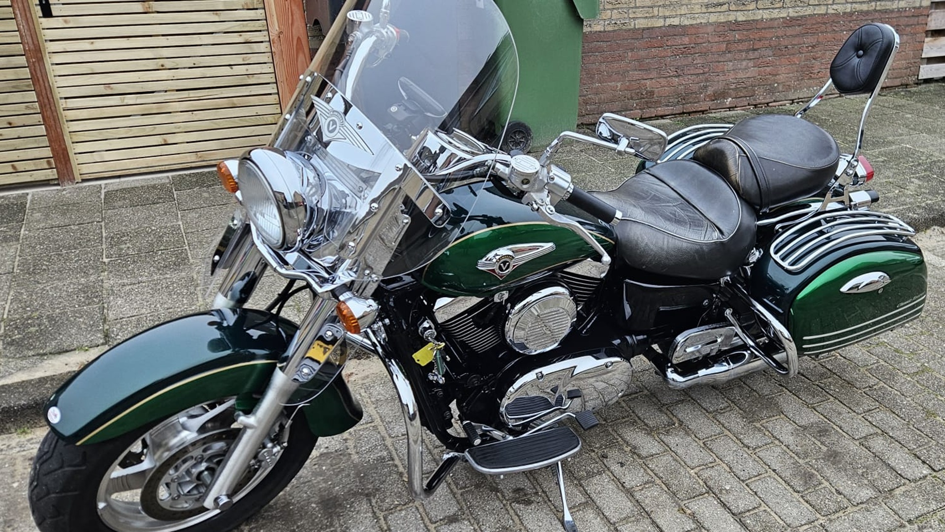 Donkergroene cruiser motorfiets met lederen zadel, glimmende chromen accenten en een hoog windscherm, geparkeerd op een stenen oprit.