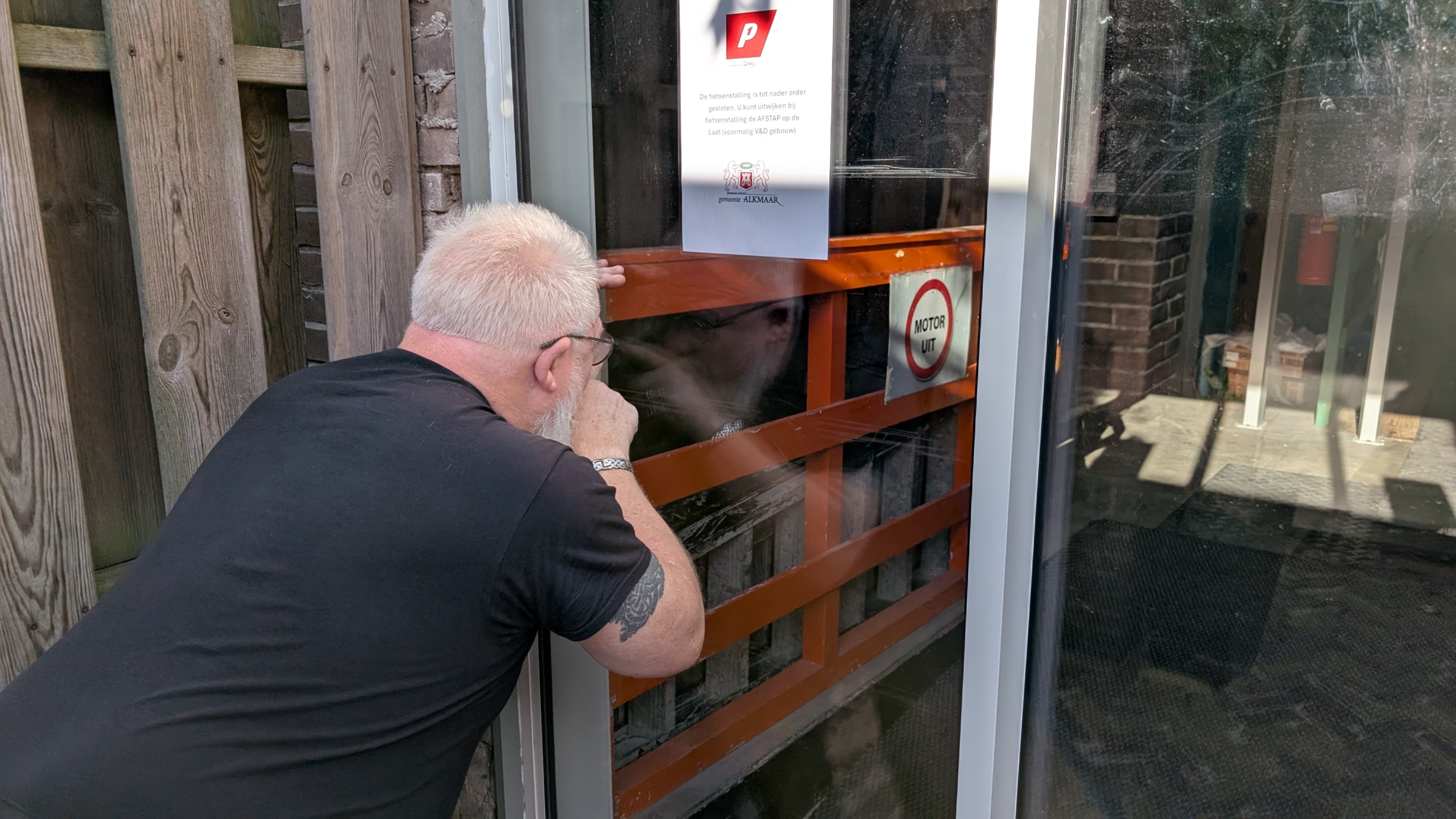 Een man met een witte baard en een zwart T-shirt kijkt door een glazen deur met ramen en een bord waarop "MOTOR UIT" staat. Er hangt ook een bordje met een mededeling van de gemeente Alkmaar.