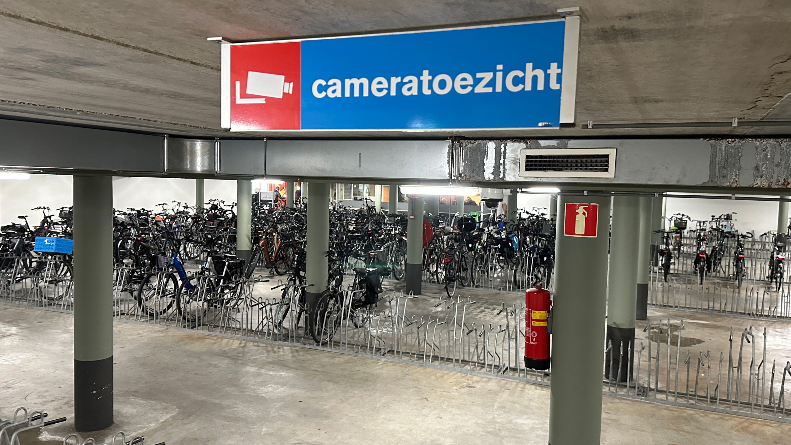 Fietsenstalling met rijen fietsenrekken en verschillende geparkeerde fietsen, met aan het plafond een bord met "cameratoezicht" en een pictogram van een camera.